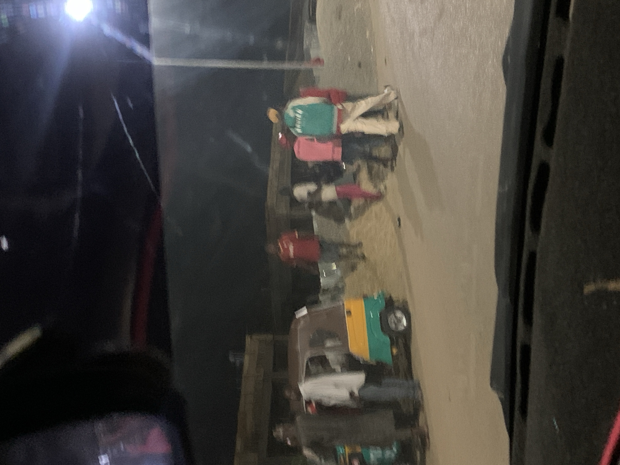 The image, seemingly taken from inside a vehicle, depicts a street scene at night in Barakin Akawo, Nigeria. On the right side of the frame is a light-colored paved road. Several individuals are present, appearing to be walking along the road. At least one auto-rickshaw, characterized by its brown, yellow, and green coloring, is visible parked alongside the road. The overall environment suggests an urban area with pedestrian and vehicular traffic.