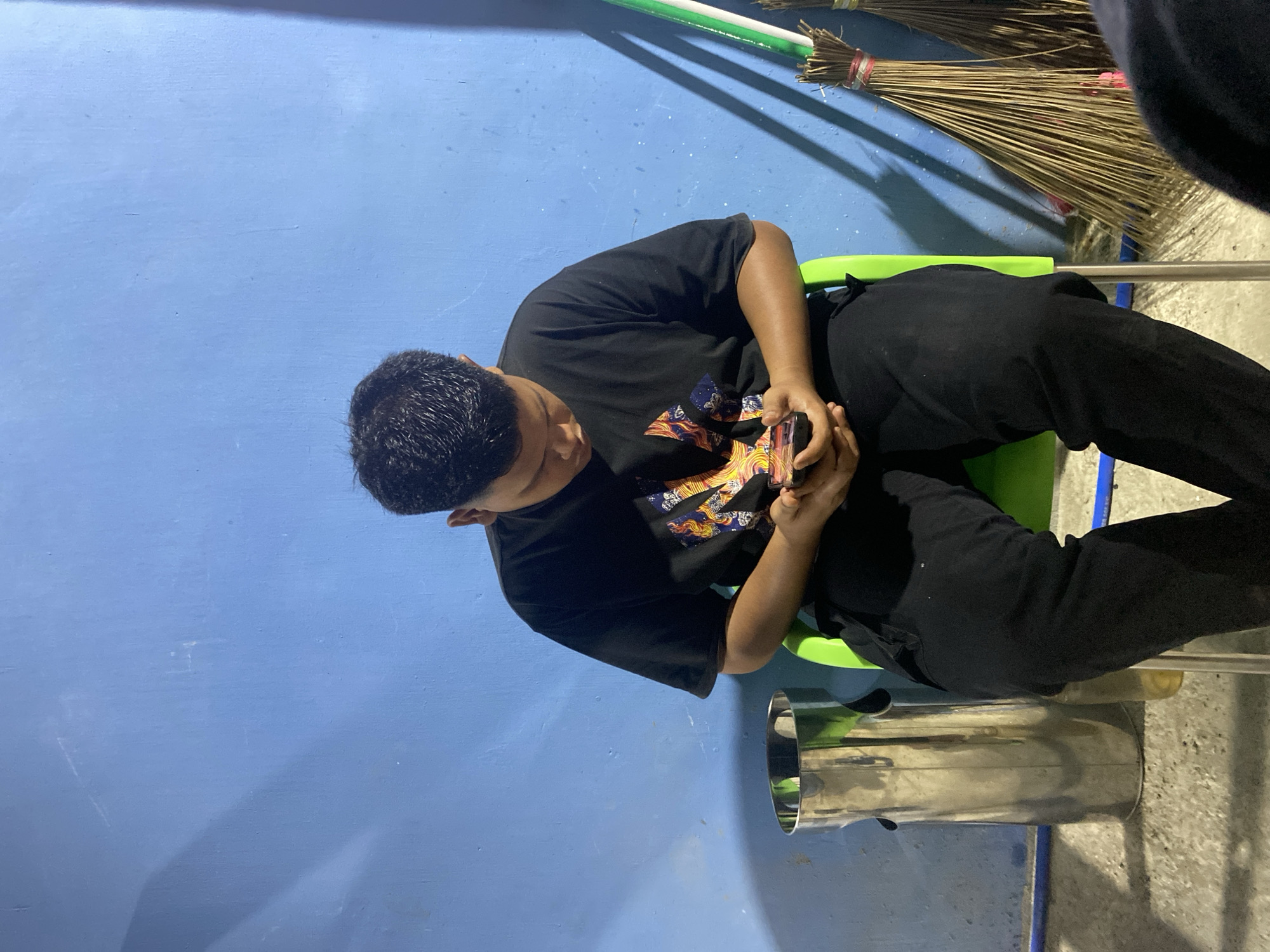An image, originally rotated 90 degrees counter-clockwise, depicts an individual absorbed in a smartphone in what appears to be an indoor or covered outdoor setting in Gampongaceh, Indonesia.
The primary subject is a person, likely male, with short dark hair, seen from an elevated perspective looking down. They are seated on a lime-green chair, wearing a black short-sleeved t-shirt with a prominent, colorful graphic design on the chest, and black pants. Both hands are holding a dark-colored smartphone, indicating they are actively using it.
The background features a distinct, textured blue wall, possibly painted concrete. To the right of the subject, several brooms made of natural fibers/straw are leaning against the wall or in a corner, casting a shadow. Adjacent to the person's right leg and the chair, a shiny, metallic cylindrical object, potentially a trash can or part of a water dispenser, is visible. The floor beneath the chair and metallic object is a grey concrete surface.
The activity shows the person focused on their device, suggesting they are browsing, texting, or playing a game. The lighting, with distinct shadows, hints at artificial illumination or a specific time of day like evening or night. No strong emotions are discernible on the subject's face, only concentration. There is no visible text in the image.