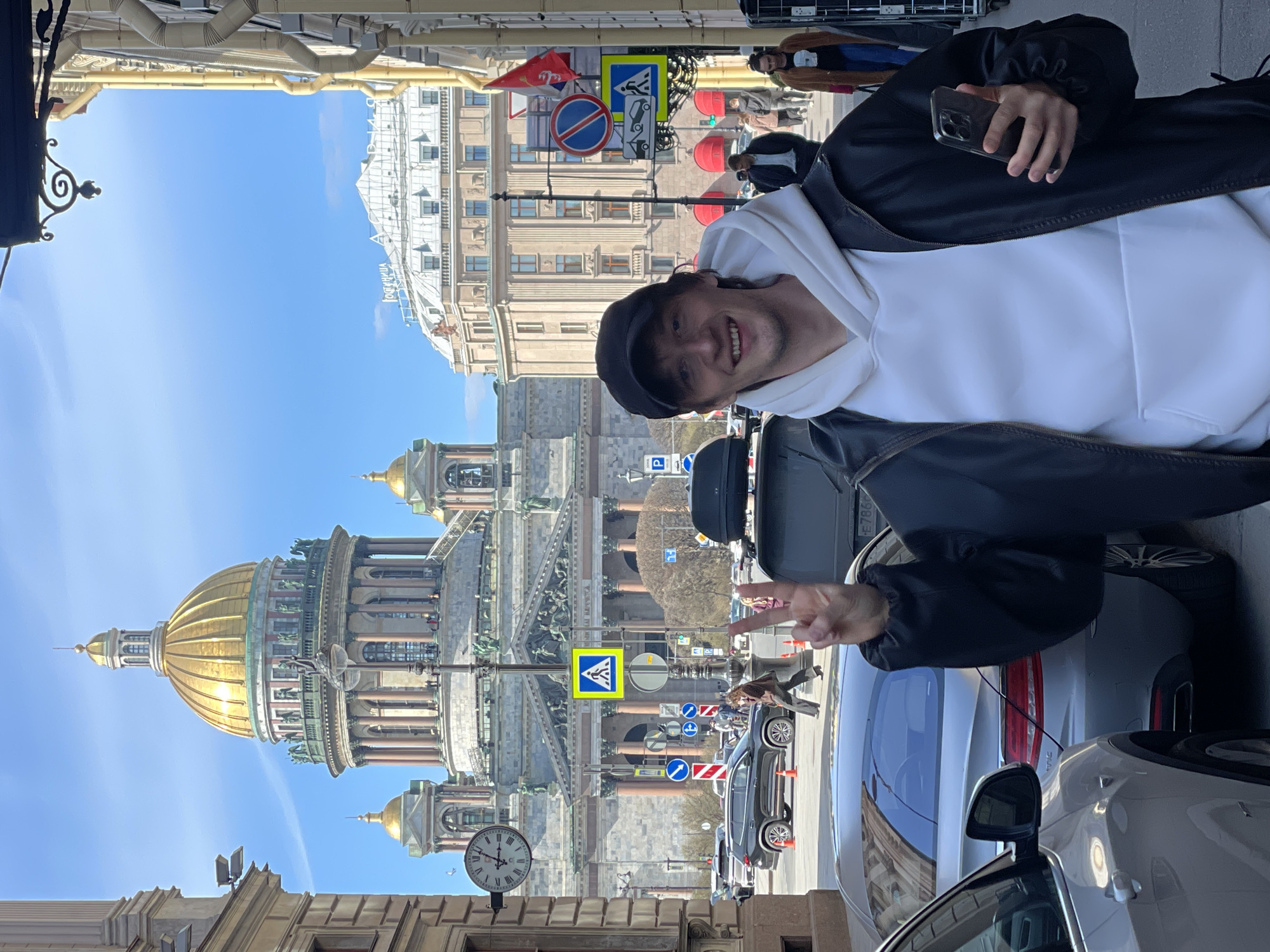 The image captures a vibrant street scene in Saint Petersburg, Russia, on a clear, sunny day. The main subject, a young man, is prominently featured in the foreground, smiling widely and making a peace sign with his left hand while holding a smartphone in his right, suggesting he is taking a selfie or being photographed. He is dressed in a black baseball cap, a white hoodie, and a black jacket.
Behind him, the magnificent golden dome and classical facade of Saint Isaac's Cathedral dominate the midground, serving as a clear architectural landmark for the city. Numerous parked cars are visible on the street in the foreground and midground. In the background, beyond the cathedral, other multi-story buildings line the street, and several people can be seen walking.
Notable details include a wall-mounted clock with Roman numerals, indicating an approximate mid-day time. Various road signs are visible, including a blue pedestrian crossing sign, a round red "no stopping" sign, and a blue parking sign. On a distant building, the mirrored Russian text "ГОСТИНИЦА" (Gostinitsa), meaning "Hotel," is discernable. The bright blue sky and ample sunlight suggest pleasant weather conditions, contributing to the cheerful emotion conveyed by the main subject.