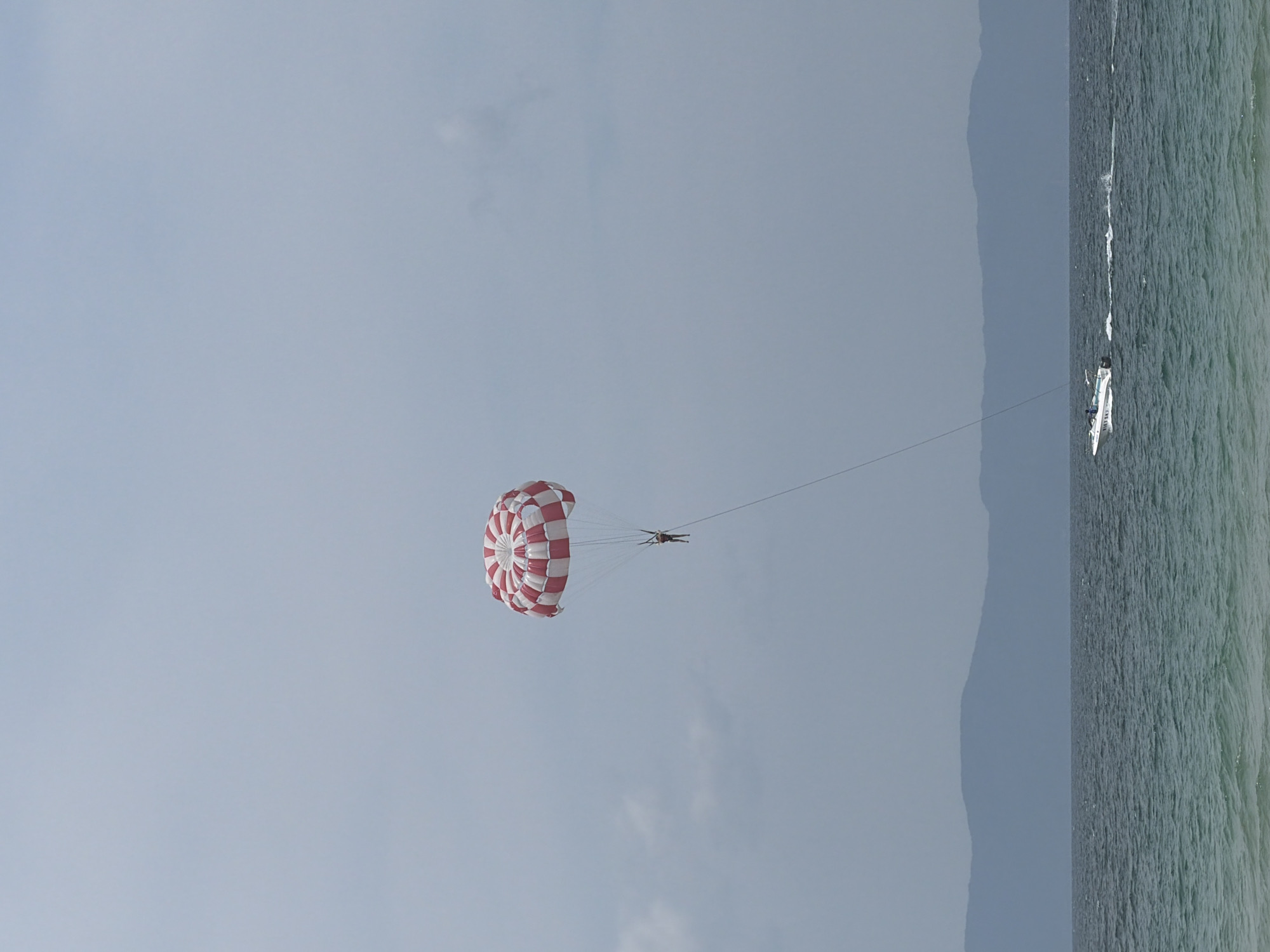The image depicts a parasailer being towed by a boat on the ocean. The parasail is red and white, and the person is suspended beneath it. The boat is white and is moving away from the viewer, leaving a wake in its path. In the background, a range of hazy blue mountains can be seen. The sky is overcast with light clouds. The water is a deep blue-green, with small waves. The overall scene suggests a recreational activity taking place at a coastal location, possibly in the city of Dong Ninh Hoa Ward, Vietnam, given the mountainous coastline.