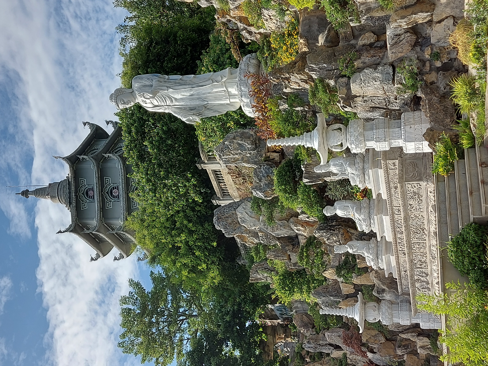The image shows the Linh Ung Pagoda in Da Nang, Vietnam, with the White Buddha statue prominently displayed. The statue of the White Buddha, a symbol of compassion, stands tall on a lotus-shaped pedestal, overlooking the city and the sea. Behind the statue, a tiered pagoda rises, adorned with intricate carvings and dragon motifs. The pagoda is surrounded by lush greenery, with rocks and smaller Buddha statues forming a serene landscape. The sky above is a clear blue, dotted with white clouds, adding to the tranquil atmosphere of the scene. The overall impression is one of peace and spiritual devotion.