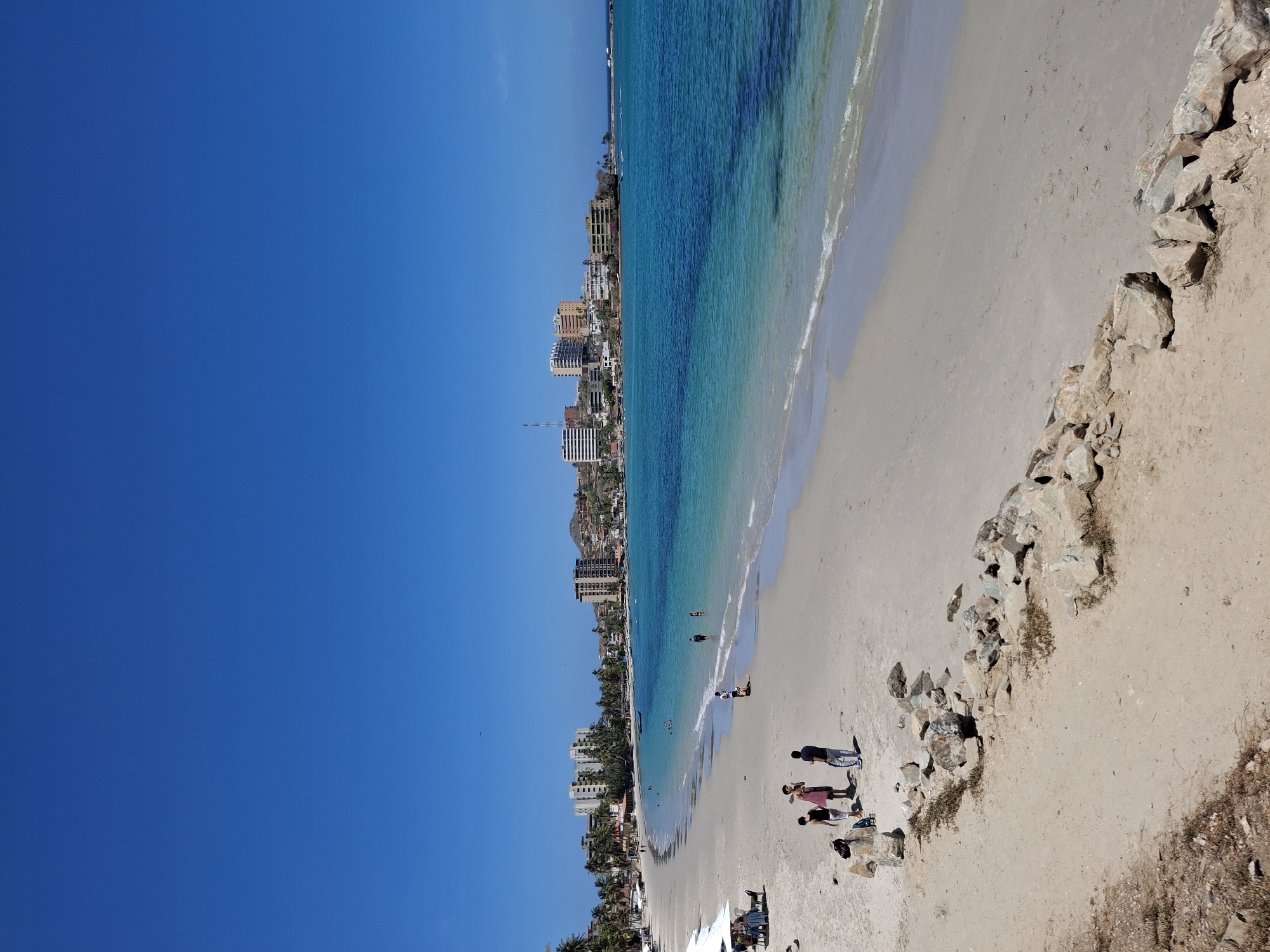 The image depicts a beach in Pampatar, Venezuela, viewed from an elevated perspective. The sky is a clear, vibrant blue. A coastline of buildings stretches along the left side of the image, juxtaposed against the beach. The turquoise-colored ocean meets the sandy shore. Several individuals are visible on the beach: some are standing in a group, while others are near the water. The shoreline is bordered by rocks and sandy terrain.
