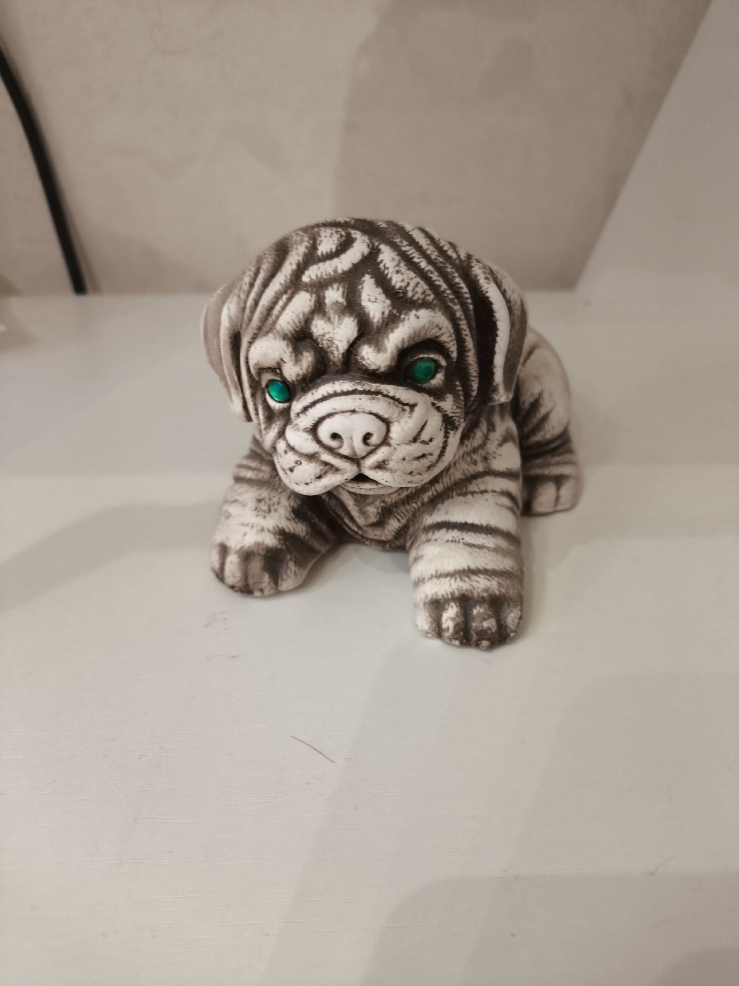 The media file displays a ceramic dog sculpture positioned centrally, appearing to lie down. The sculpture depicts a brachycephalic dog breed, possibly a pug, with a distinctive patterned coat featuring dark brown or grey stripes and swirls over a lighter, off-white base, giving it a somewhat stylized, tiger-like or marbled appearance. Its eyes are a striking, vibrant green, contrasting sharply with the monochromatic body.
The dog figure is presented in a resting pose, with its front paws extended forward. The background is simple and light-colored, suggesting an indoor setting like a shelf or table against a wall. A faint dark vertical line or object is partially visible on the far left of the frame. The lighting is soft and even. No text is visible in the image. This scene is located in Khimki, Russia.