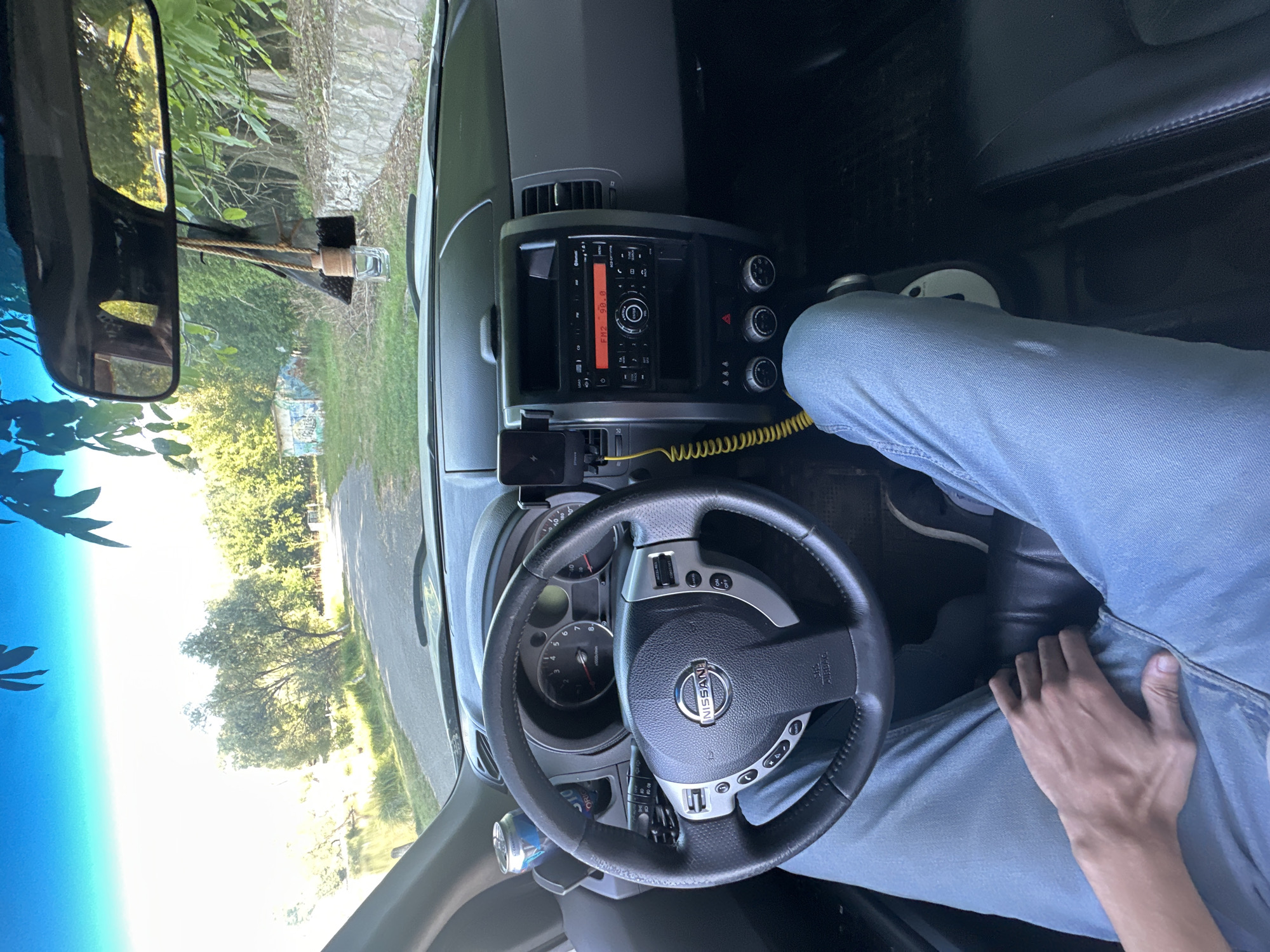 The image shows the interior of a Nissan vehicle parked on a dirt road next to a body of water, likely a river or lake, with trees and vegetation on either side. In the background, a graffitied building and a stone wall are visible. The car's dashboard, steering wheel, and center console are prominently displayed, with a phone mount and a can of soda on the passenger side. The driver's legs, clad in light blue jeans, are visible in the foreground, with their hand resting on their lap. The scene suggests a stop during a road trip or a moment of relaxation in a natural setting. The location is identified as Makiv, Ukraine.