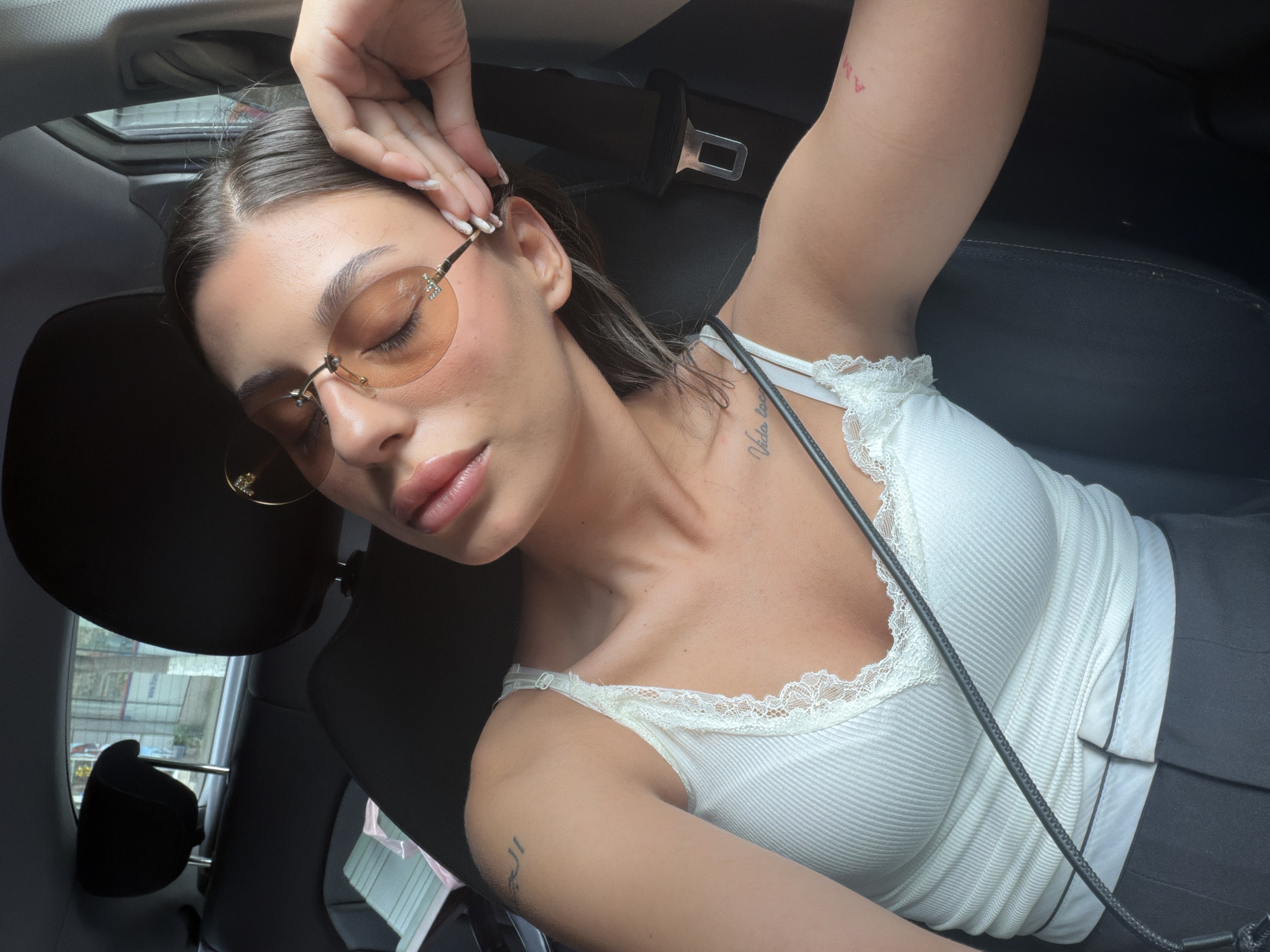 The image shows a woman in the passenger seat of a car. She is wearing a white lace-trimmed tank top and sunglasses with orange lenses. Her eyes are closed, and her hand is resting on her face. The interior of the car, including the headrest, seatbelt, and dark upholstery, is visible. Outside the car window, a blurred city scene can be seen, suggesting an urban environment. There are no specific landmarks or indicators that would place the scene in Burgas, Bulgaria.