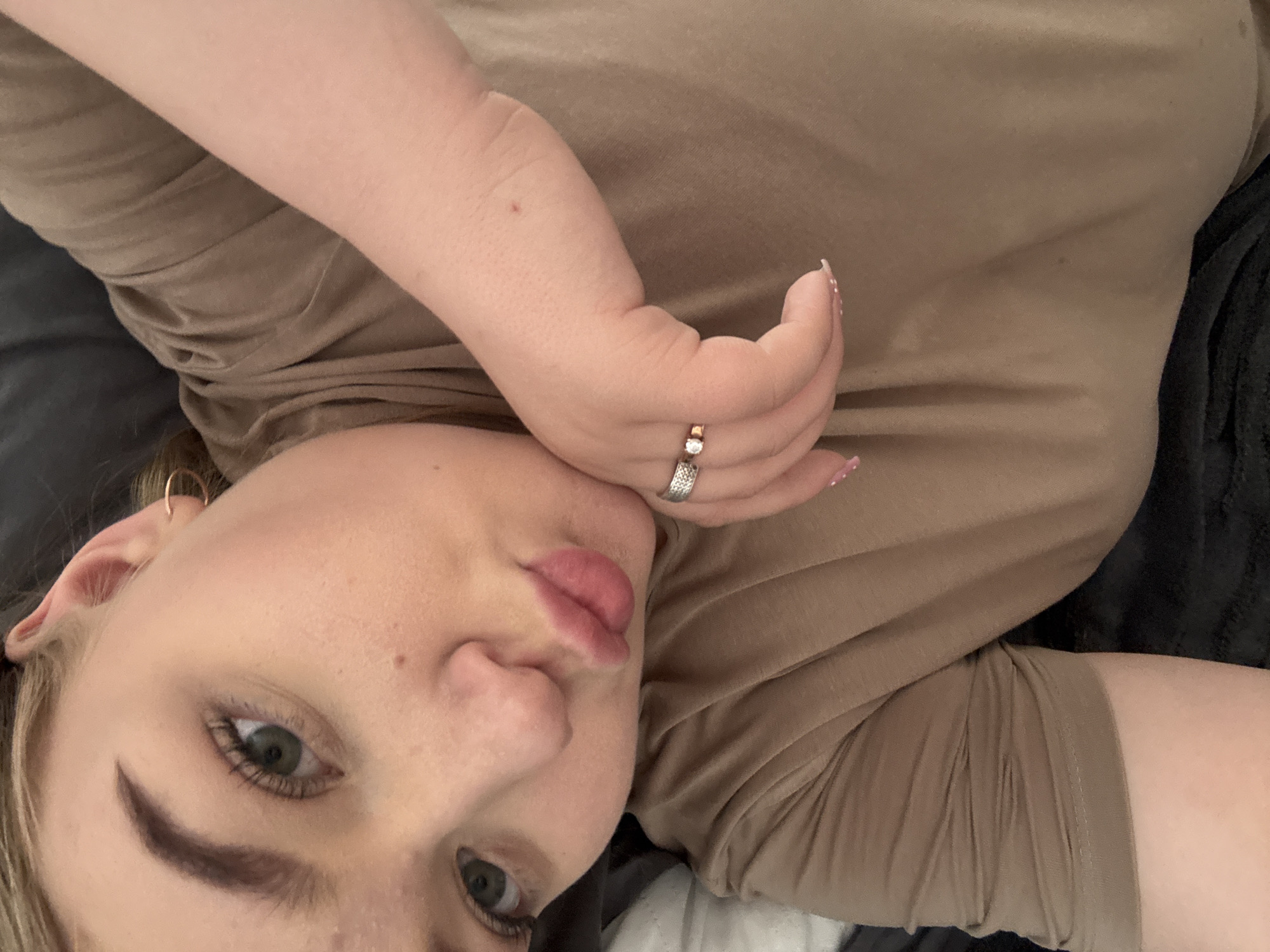 A woman is lying down, likely indoors, wearing a brown shirt and a silver ring. Her left hand is near her face. The background consists of a dark surface. The location is unknown but presumed to be Chaikovsky, Russia.