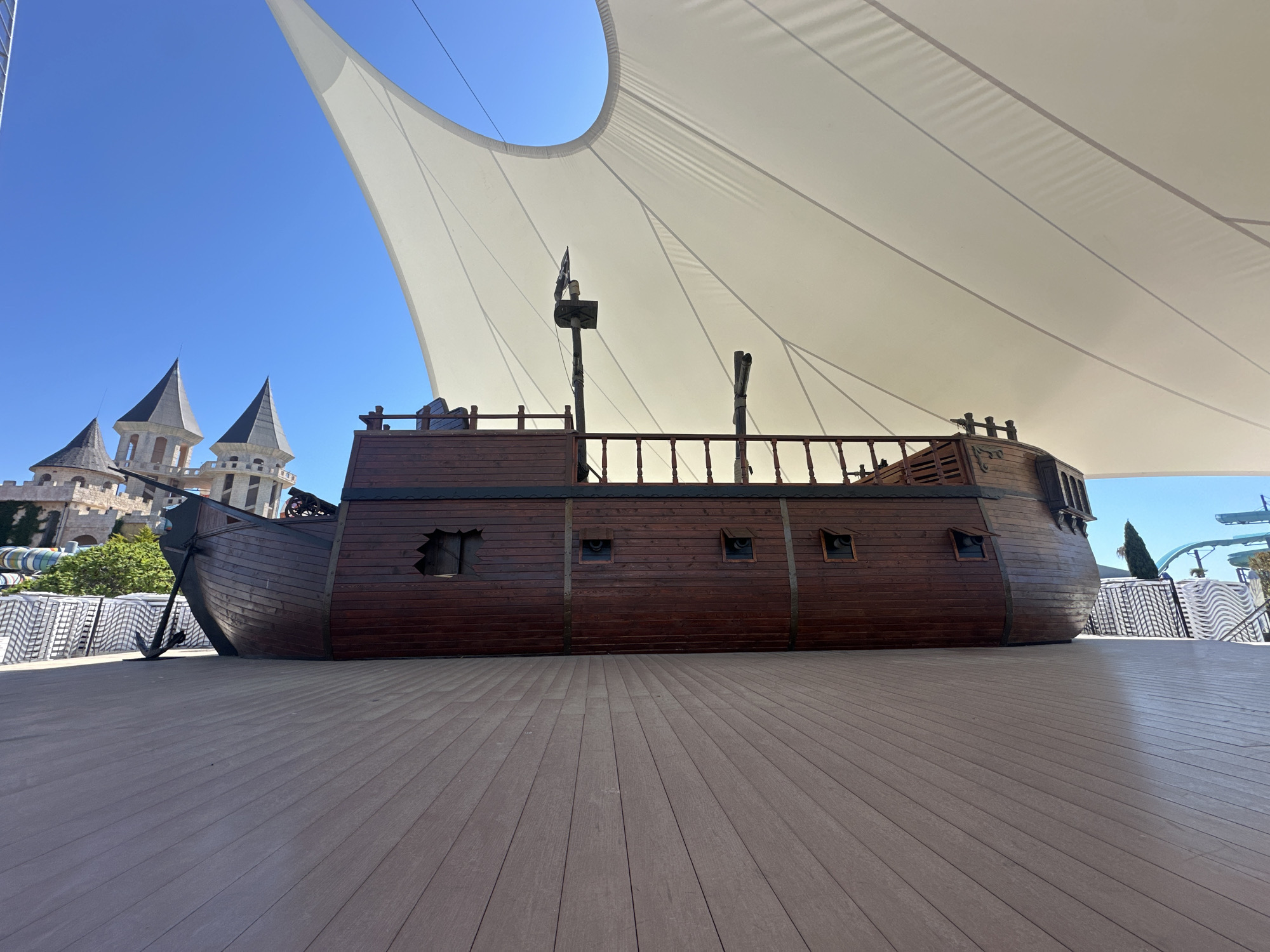 The image captures a large, static, wooden pirate ship replica situated on a wide, wooden deck under a vast, white, tensile fabric canopy. The ship is dark brown, features a railing, a broken "window," and other faux details like cannon ports and a mast. An anchor hangs from the front of the ship. The deck is composed of horizontally aligned planks that converge in the distance. In the background, partly obscured by the ship and canopy, are castle-like structures with pointed towers, potentially part of a theme park or resort. Stacks of white lounge chairs are visible to the right of the ship. The sky is clear and blue. The location is Ravda, Bulgaria.