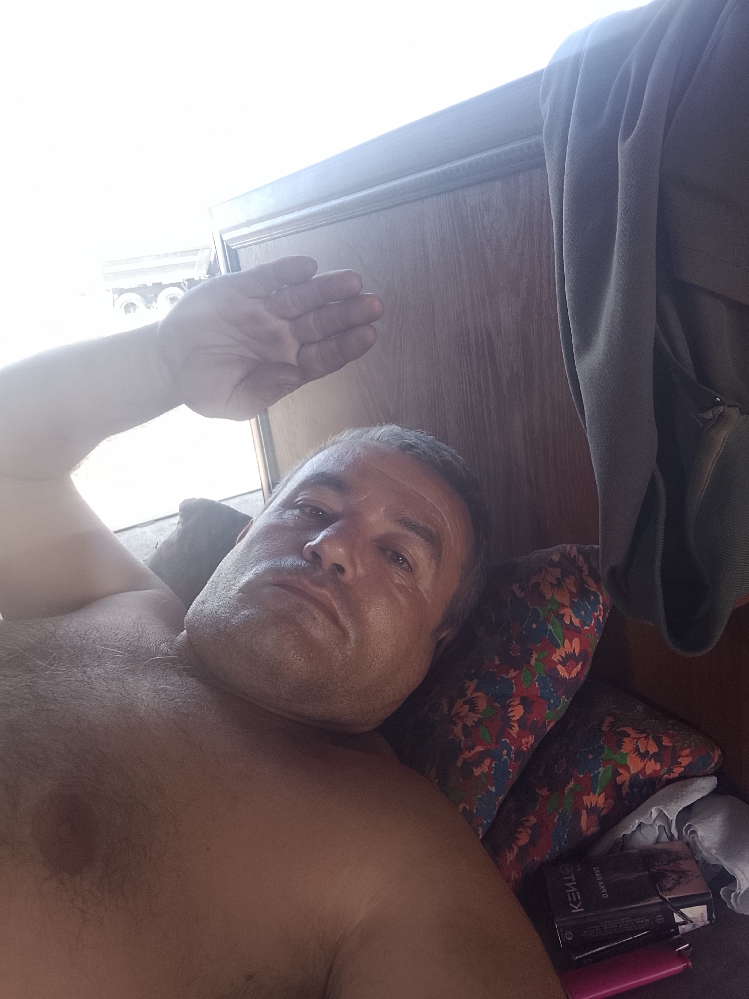 A medium shot captures a fair-skinned man lying on his back on a pillow with a floral pattern. He is shirtless, revealing his chest and shoulder. His left arm is raised, with his hand shielding his face from bright light originating from the upper left of the frame. His head is turned slightly to the right, and he appears to be looking upwards. Behind him, to the right, a dark grey garment is draped over a wooden surface. In the lower right corner, a cigarette pack and a pink lighter are visible. The background shows a bright, washed-out area with a hint of a vehicle, possibly a truck, and a wooden panel. The overall lighting suggests an outdoor setting or a room with ample natural light. The location is identified as Sabir Rakhimov, Uzbekistan.
