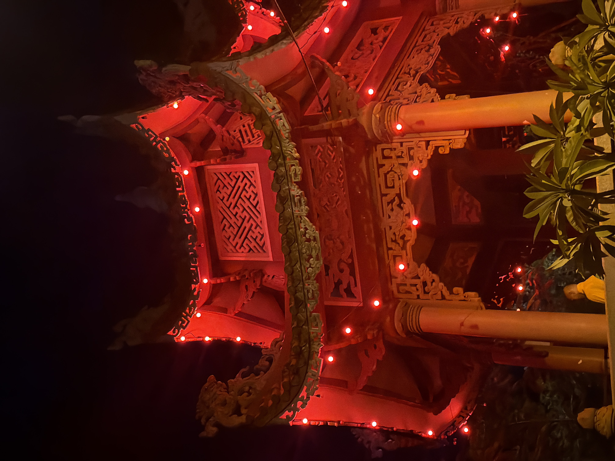 The image depicts the intricately designed roof and upper structure of a traditional East Asian pavilion, illuminated by numerous small red lights. The pavilion is made of wood and features ornate carvings, decorative eaves, and multiple tiered roofs, a common architectural style in East Asian countries. The color palette is dominated by the vibrant red of the lights and the structure itself, contrasting with the dark night sky. In the foreground, lush green foliage is visible, suggesting the pavilion is set in a garden or park. To the lower right, a person wearing a yellow robe, possibly a monk, is partially visible, adding a human element to the scene. The overall impression is one of a beautifully lit architectural monument at night. This pavilion is part of the Long Son Pagoda complex in Nha Trang, Vietnam, a significant Buddhist temple known for its large white Buddha statue.