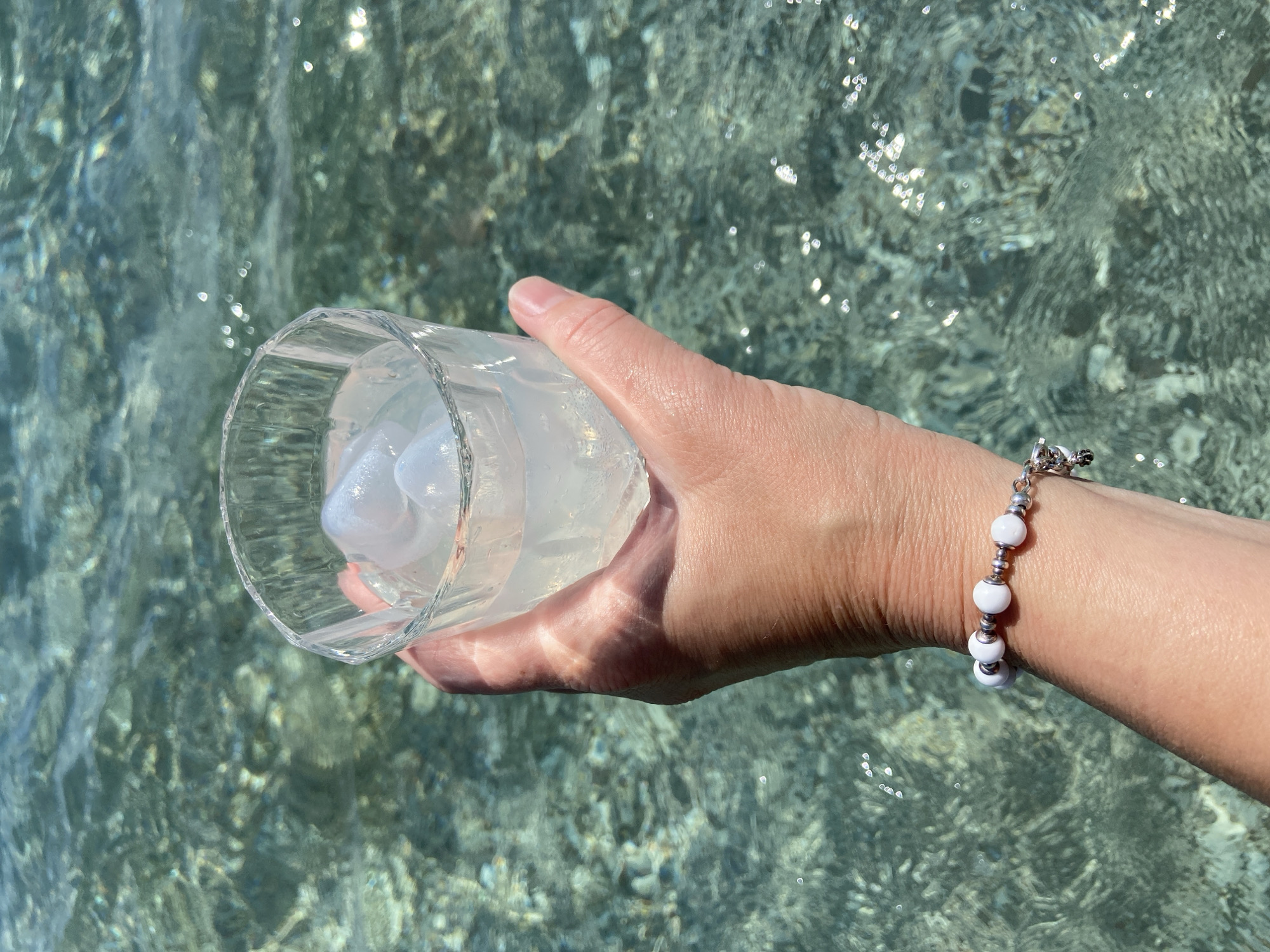 A human hand holds a clear glass filled with liquid and white solids. The background shows clear, shallow water with sunlight reflecting on its surface. A bracelet of white and silver beads adorns the wrist. The location appears to be coastal, likely in Budva, Montenegro, given the water's clarity and the prevalence of such scenes in the region. No other people or specific objects are discernible.
