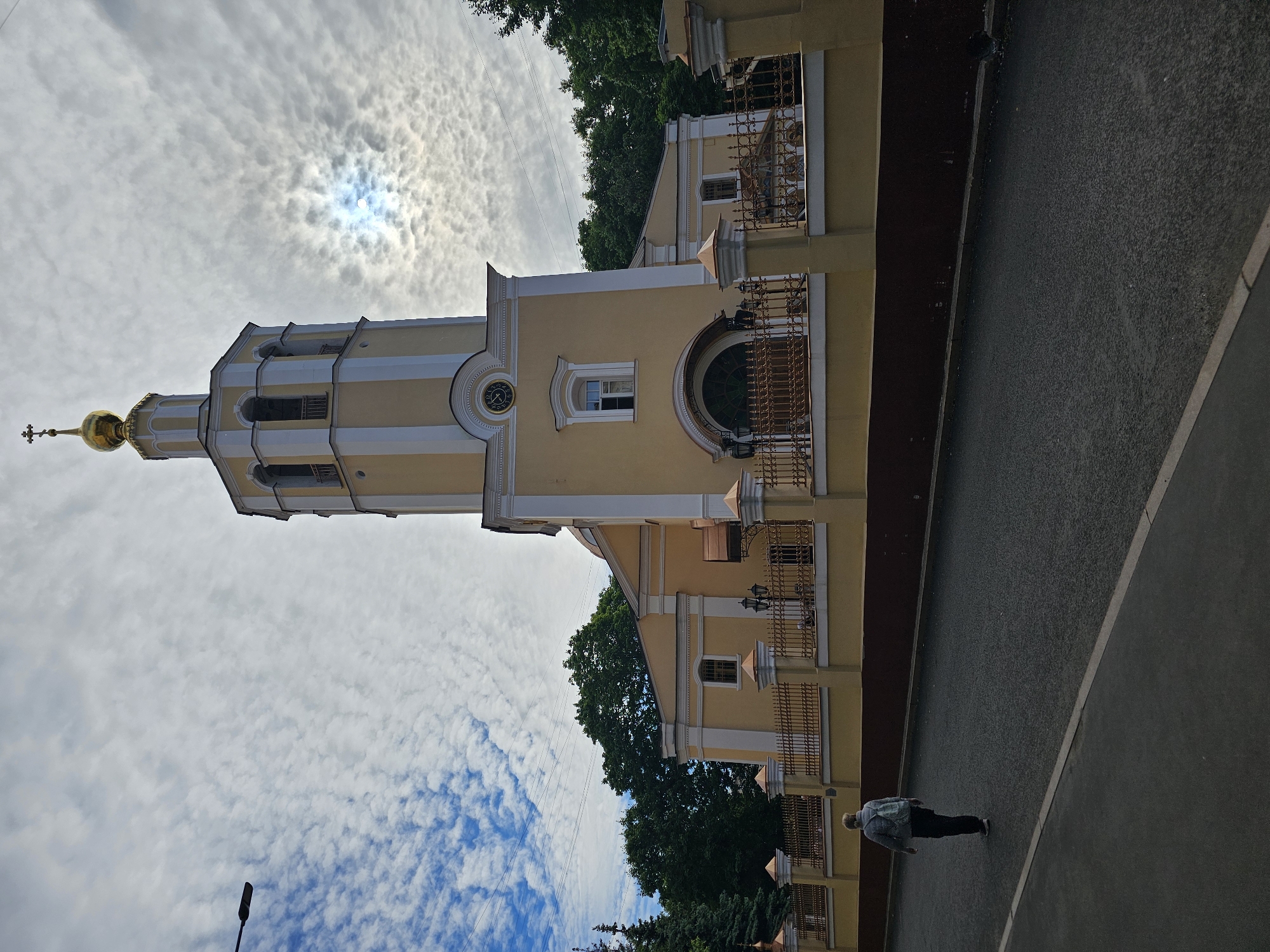 The image shows the bell tower of the Church of the Assumption of the Blessed Virgin in Khimki, Russia. The tower is yellow with white accents and a golden dome topped with a cross. A person is walking on a paved area next to the church. The sky is cloudy with the sun visible through the clouds. Trees surround the church building.