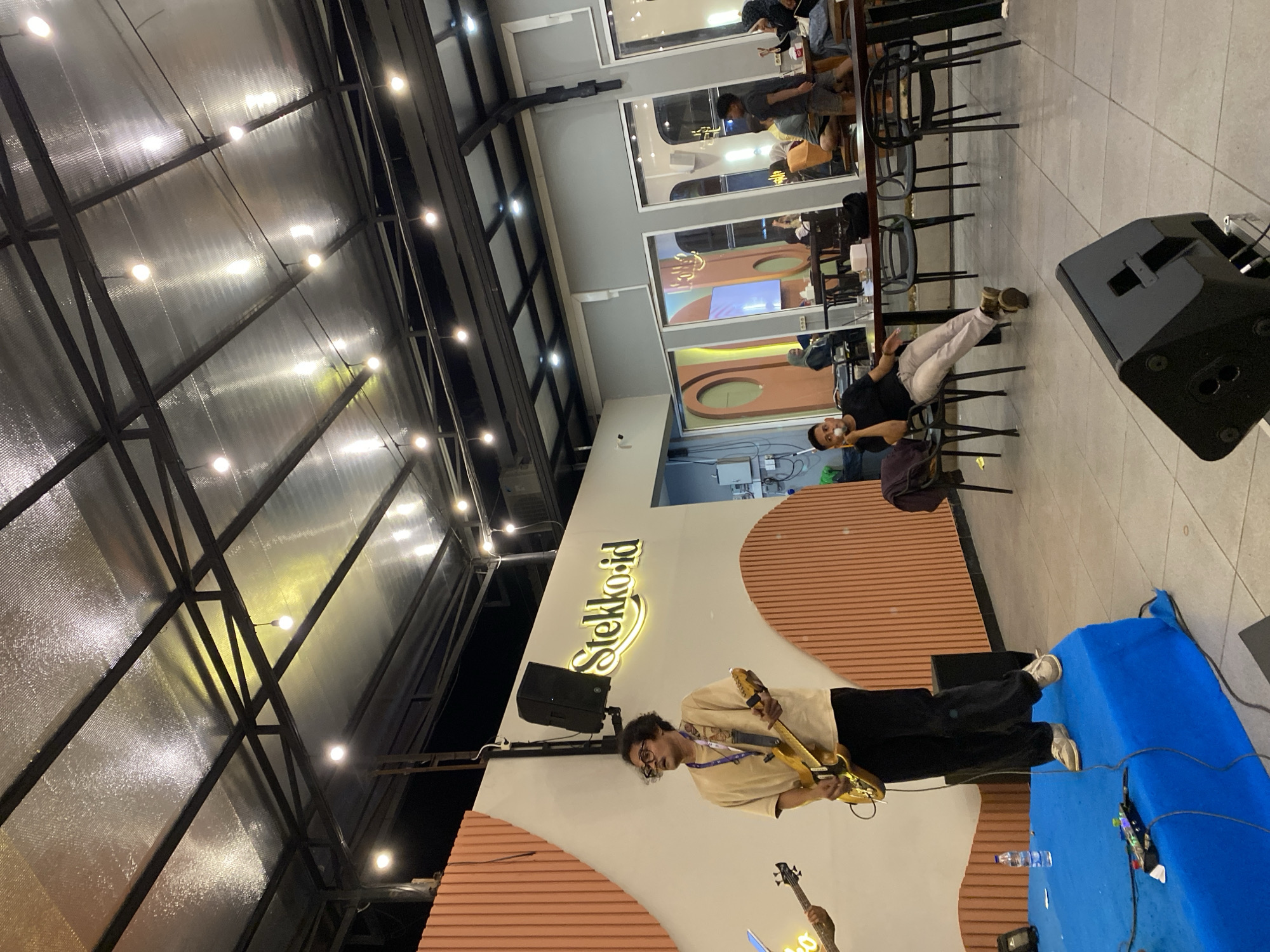 The image shows a musical performance in an indoor setting in Lhokseumawe, Indonesia, as indicated by the "stekko.id" signage. A man is playing an electric guitar while standing on a blue-covered platform. To the right of the stage, a black speaker is visible. Several individuals are seated at tables in the background, some appearing to observe the performance. The building has a high ceiling with exposed metal beams and numerous small lights. The interior design includes peach-colored striped patterns on the walls and arched windows. Chairs and tables are arranged in rows along the side of the room. A water bottle is visible on the stage near the musician.