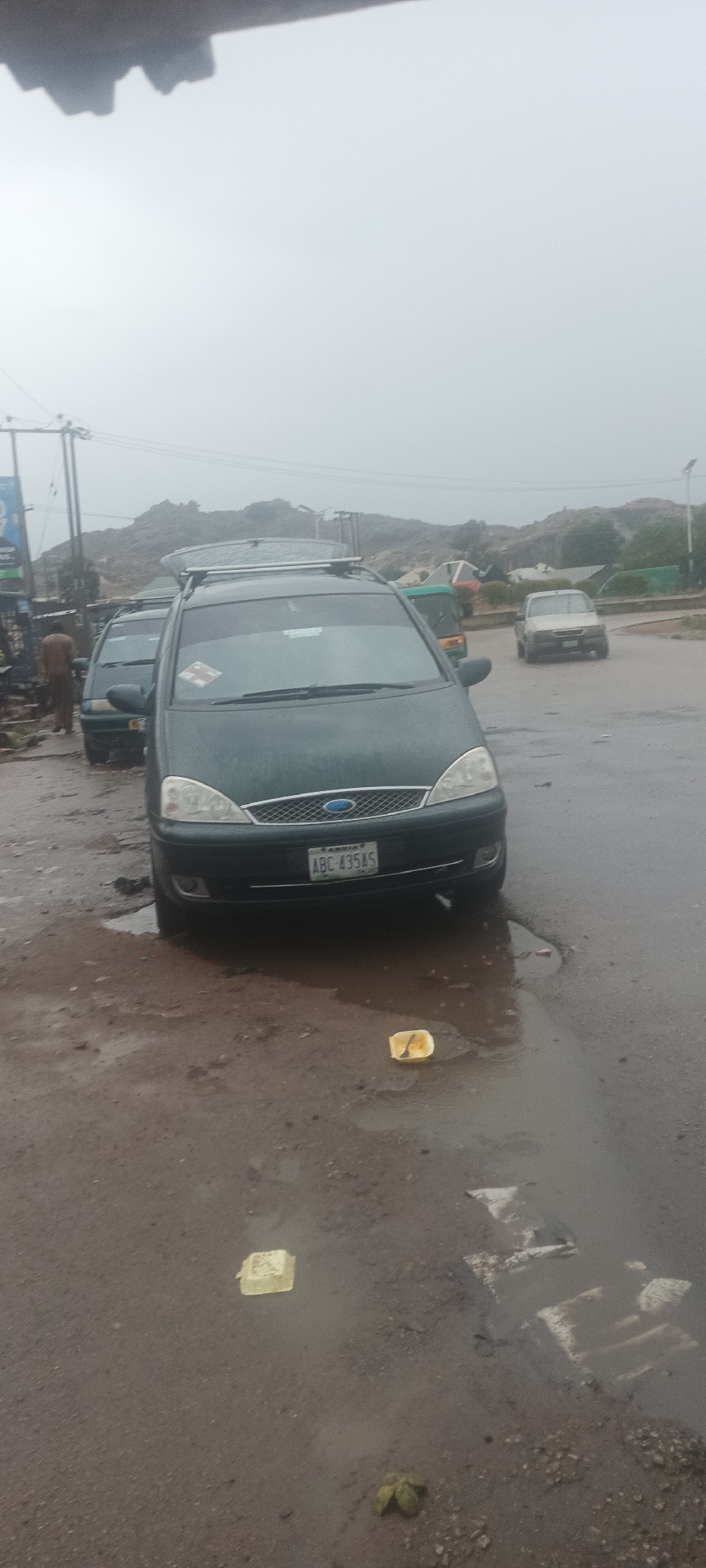 The image shows a wet, overcast street scene in Kuru Dungus, Nigeria. A dark green Ford Galaxy minivan with roof rack is parked on the side of the road, partially in a puddle. The license plate reads "ABC 435AS". The road surface is cracked and potholed. Scattered litter, including yellow foam containers, is visible in the foreground. A man wearing dark clothing stands near the minivan on the left side of the frame. In the background, other vehicles are present, including a light-colored car approaching an intersection. Buildings and rock formations are visible in the distance. Power lines are strung above.