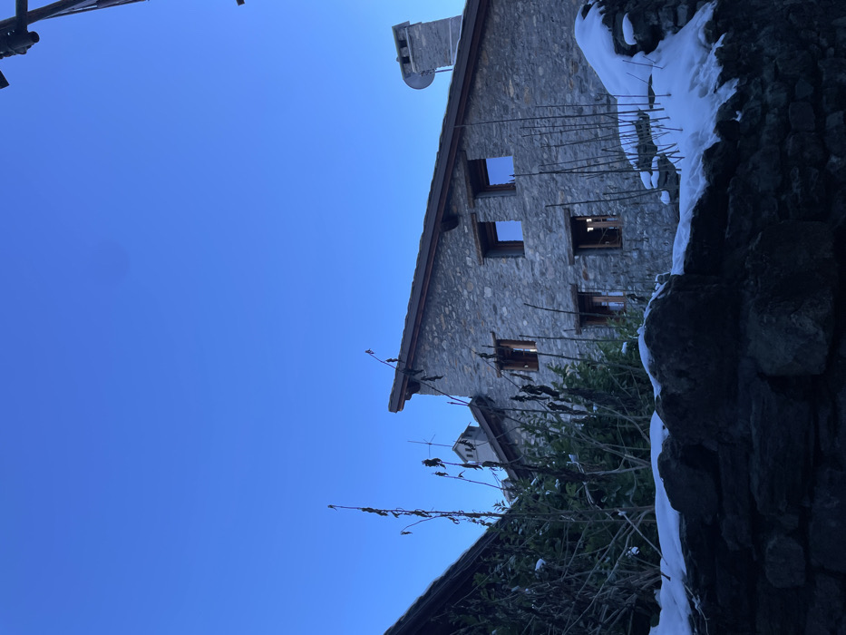 The photograph, when viewed in its intended landscape orientation (rotated 90 degrees clockwise from the uploaded version), depicts a traditional stone building under a clear, bright blue sky in a snowy, winter setting in Courmayeur, Italy.
The main subject is a rustic, multi-story building constructed from rough, irregular grey and brown stones, featuring a gabled roof made of dark material. A stone chimney extends from the roofline, and a small, partially visible satellite dish is mounted near it. Several rectangular windows are visible on the facade; some appear as empty frames or reveal dark interiors, suggesting the building might be unoccupied or seen from a distance.
In the immediate foreground, along the right side and bottom of the frame, is a rugged stone wall thickly covered with white snow. Scattered bare, dark tree branches and some evergreen bushes, also lightly dusted with snow, fill the lower and mid-ground, partially obscuring the base of the building.
The sky is a vibrant, cloudless blue, indicating a clear and sunny daytime. The presence of abundant snow and bare branches signifies cold winter conditions. There are no people or explicit activities taking place, contributing to a tranquil and serene atmosphere. No text is visible within the image. The scene captures the quiet beauty of an alpine winter day.
