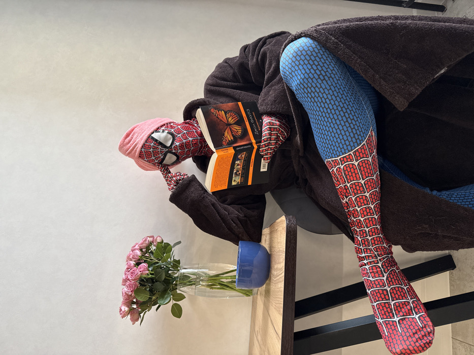An individual dressed in a red and blue Spider-Man costume, a dark brown bathrobe, and a pink towel wrapped around the head, sits in a chair in an indoor setting in Kazan, Russia. The person is holding an open book with an orange cover featuring a butterfly design and Russian text identifying "JOHN FOWLES" as the author and "THE COLLECTOR" as the title. The right index finger is pressed against the temple of the Spider-Man mask. One leg, visible from the knee down in the Spider-Man suit, is extended forward. On a wooden table to the left, a clear glass vase contains a bouquet of numerous small pink roses, and a blue ceramic bowl is present. The background is a plain, light-colored wall.