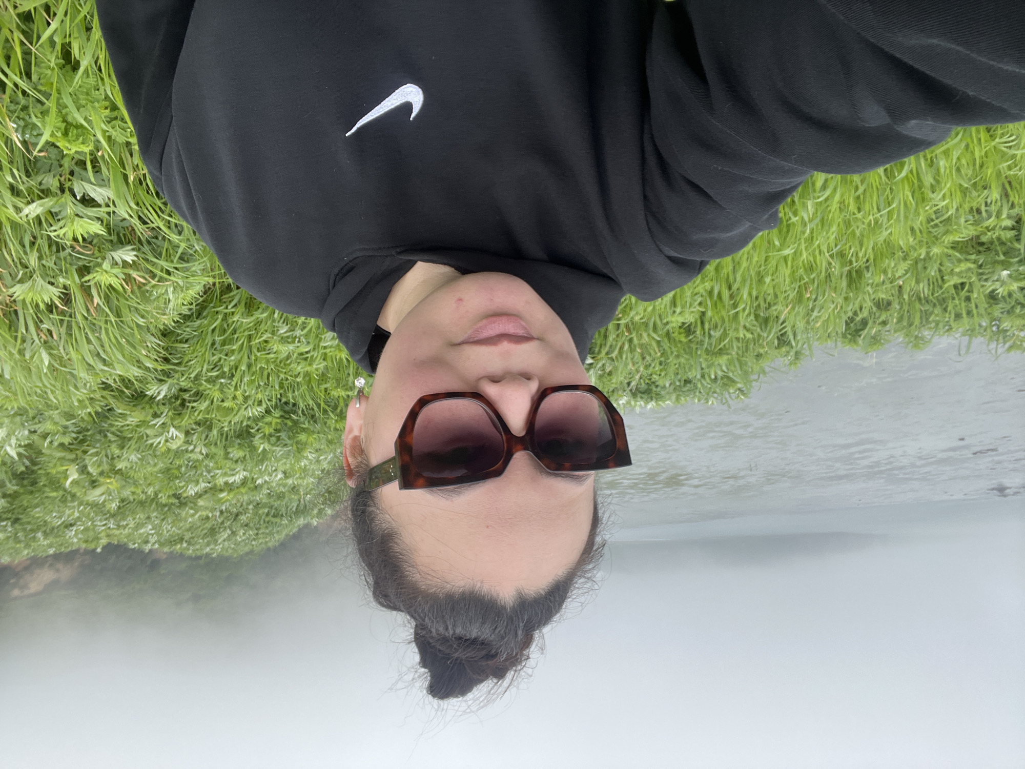 A woman is taking a selfie on a foggy day in Tikhoe, Russia. She is wearing a black Nike hoodie and large, tortoiseshell sunglasses. Her dark hair is pulled up into a bun. Behind her, the scene is dominated by lush green grass, with a body of water and a distant, fog-shrouded landmass visible in the background.