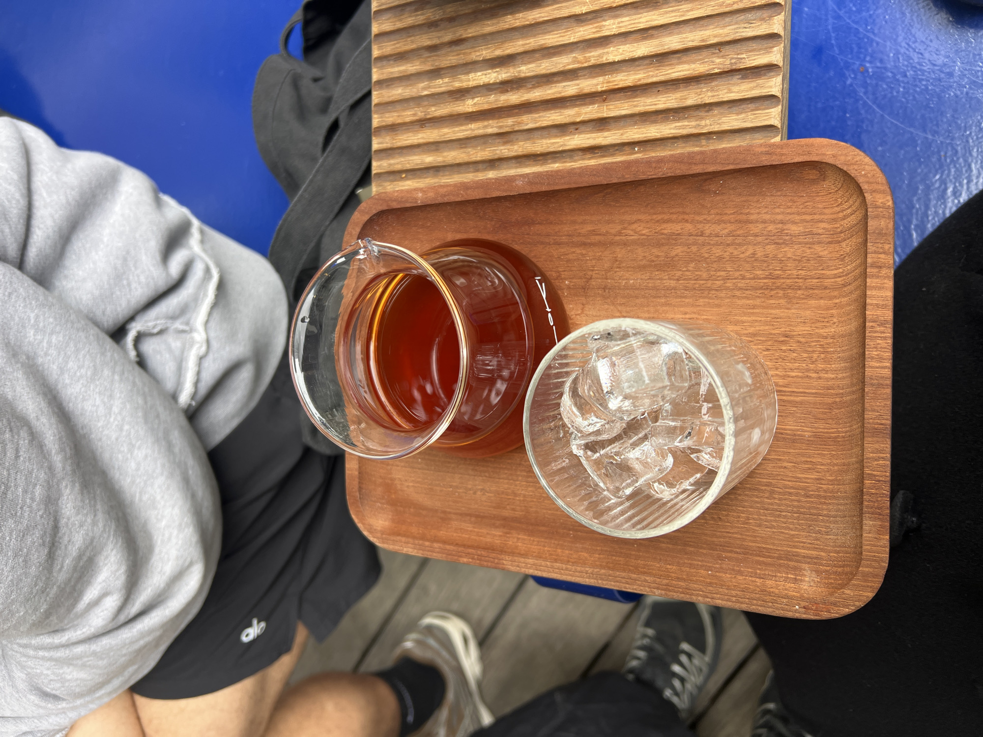 The image provides an elevated, close-up view of a beverage service in Tel-Aviv, Israel. A person, partially visible, wears a grey hooded sweatshirt and black athletic shorts with an "alo" logo. Their left leg, featuring a beige sneaker with a white sole and a dark sock, is also visible. On a light brown wooden serving tray with rounded corners, a clear glass carafe, approximately half-full of dark amber liquid (consistent with coffee or tea), displays a visible "-Lovi" measurement mark. Alongside it, a clear glass tumbler is filled with multiple ice cubes. To the right of this main tray, a second wooden item with parallel ridges is partially visible. These items rest on a distinct dark blue surface, likely a table, which also holds a partial view of a dark fabric bag. The flooring beneath the seated individual consists of light-colored wooden planks. No specific human action is depicted, but the setup suggests preparation for beverage consumption in a casual environment.