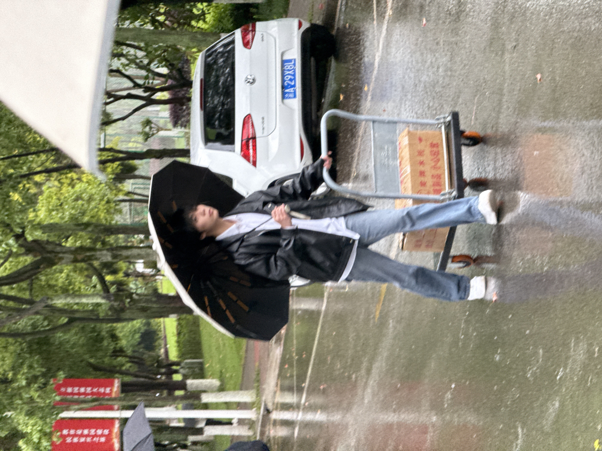 The image captures a rainy outdoor scene, likely on a paved street or pathway in Jiangjin, China. The primary subject is a young individual, possibly male, dressed in a black jacket, white t-shirt, blue jeans, and white sneakers, holding a black umbrella to shield from the rain. They appear to be walking or standing next to a silver hand cart carrying brown cardboard boxes. The person's gaze is directed slightly downwards, partially obscured by the umbrella.
In the background, a white Volkswagen Golf car is parked on the wet pavement. Its license plate, "渝A·29X8L", is clearly visible, indicating it is registered in Chongqing municipality (where Jiangjin is located). Lush green trees and foliage line the background, suggesting a park-like or tree-lined street setting. The ground is wet and reflects light, indicating ongoing or recent rainfall. Another partially visible umbrella belonging to another person can be seen in the far background on the left. Several red signs with unreadable Chinese characters are visible among the trees on the left side of the frame. The overall atmosphere is overcast, consistent with rainy weather during daytime.