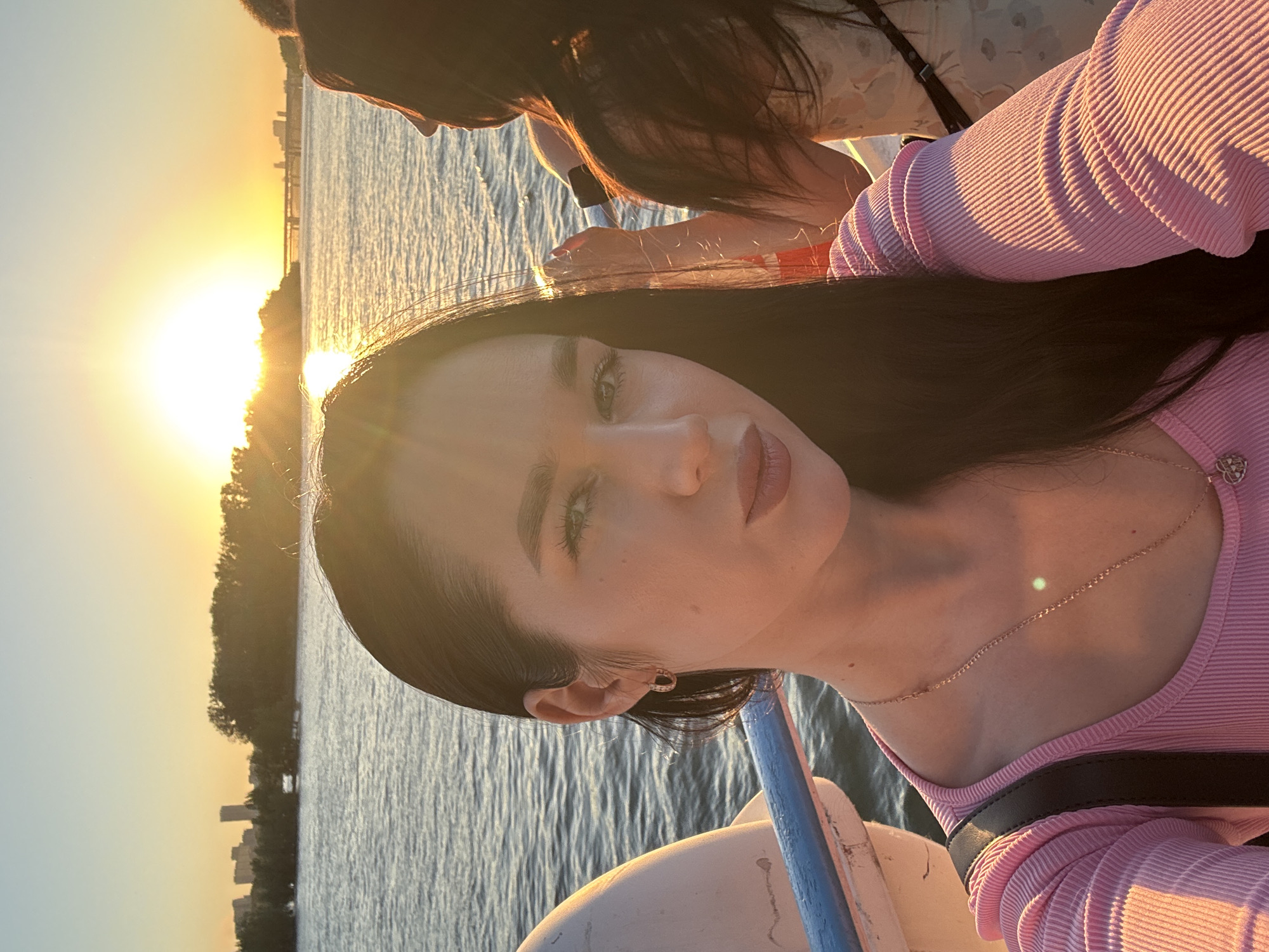 A woman with long dark hair, wearing a pink ribbed shirt and a delicate necklace, is taking a selfie on a boat. She is looking directly at the camera with a neutral expression, her head and shoulders filling the foreground. To her right, another woman with long dark hair is visible from the side, wearing a floral dress. They are on a body of water, likely a river, with a city skyline and a bridge in the background. The sun is setting, casting a warm golden glow over the scene, reflecting on the water. The water appears calm with gentle ripples. This scene is set during golden hour, creating a serene and picturesque atmosphere, likely near the Novosibirsk city embankment, Russia.