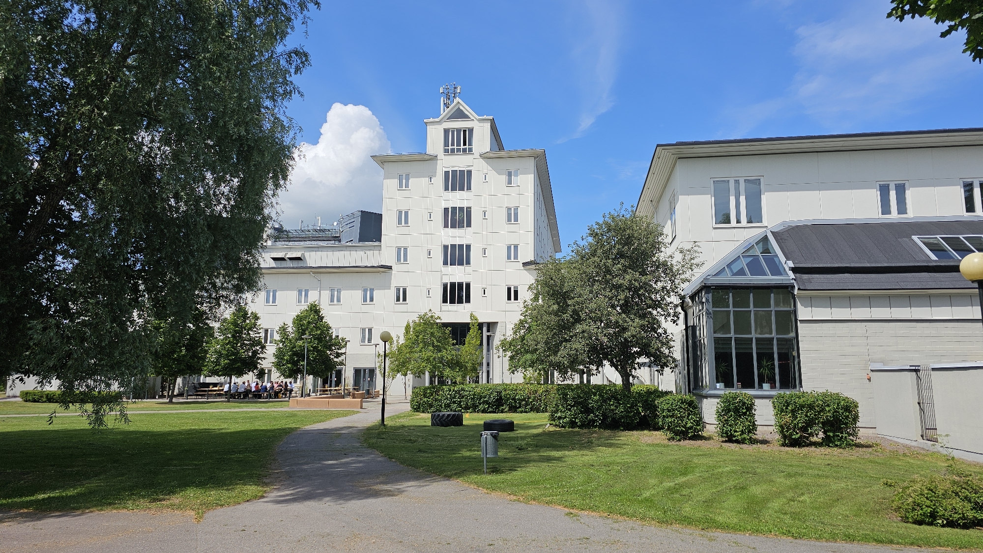 A wide-angle, eye-level outdoor shot depicts a bright, sunny day with a clear blue sky and scattered white clouds. The scene is set in the city of Linköping, Sweden, and features modern white buildings with numerous windows. A large, multi-story building dominates the center-left of the frame, its facade characterized by a series of evenly spaced rectangular windows and some taller, more complex window arrangements towards the top. To the right, another white building is partially visible, with a prominent glass-enclosed section and a dark, sloped roof.
The foreground shows a paved path curving from the bottom left towards the right, bordered by a well-maintained green lawn. Several mature trees with lush green foliage are scattered throughout the scene, with a large tree on the left casting a shadow on the lawn. In the middle ground, a group of people are seated at picnic tables under the shade of trees, appearing to be engaged in conversation or a meal. Two large black tires are placed on the grass near a small grey trash can.
The overall impression is one of a pleasant, possibly institutional or corporate campus setting, with a focus on modern architecture and green spaces.