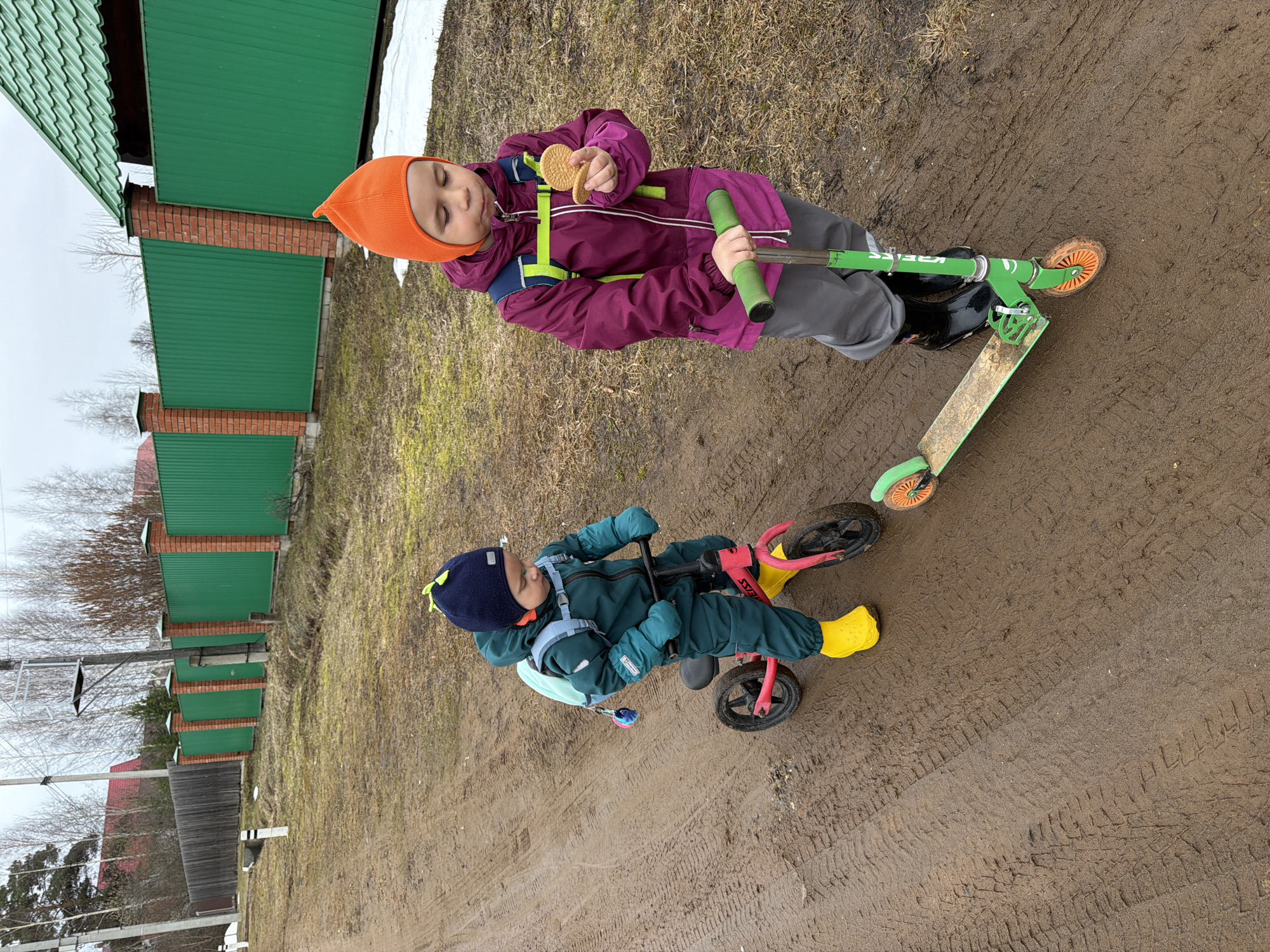 Two children are outdoors on a muddy path in what appears to be Chaikovsky, Russia. One child, wearing an orange hat, a maroon jacket, grey pants, and black boots, stands on a green scooter and holds two cookies. The other child, wearing a dark blue hat, a teal jumpsuit, and yellow boots, is on a red balance bike. A green fence and bare trees are visible in the background, suggesting a rural or suburban setting. Tire tracks and footprints are evident in the mud.