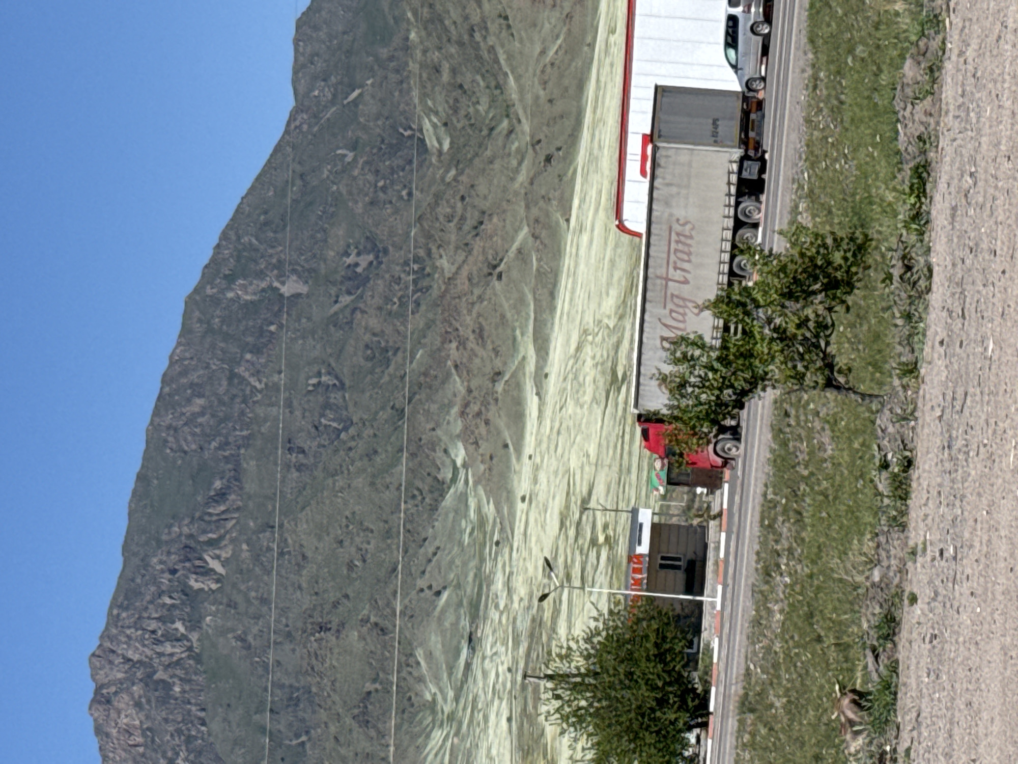 This image depicts a mountainous road scene, likely in the vicinity of Chym-Korgon, Kyrgyzstan, under a clear blue sky. A large, rugged mountain face dominates the background on the left, covered in sparse, greenish-brown vegetation and streaked with lighter, possibly dry or silty, patches.
In the mid-ground, a paved road stretches horizontally, with several vehicles, primarily large trucks, lined up or parked. A prominent white semi-trailer truck with a red cab features the text "Mag Trans" in a reddish-brown font on its trailer. Another similar truck is visible behind it, and a smaller, white delivery-type truck or van is further along the road.
Alongside the road on the right, there's a strip of grassy terrain and a rougher, lighter-colored shoulder or dirt path. A small building with a red roof and white walls, possibly a checkpoint or service station, is situated next to the road. Streetlights are also visible. Small trees with green foliage are scattered along the roadside, one appearing prominently in the foreground on the right. The scene is brightly lit by the sun, indicating daytime. No individuals are visible. The overall impression is of a transport corridor through a scenic, high-altitude landscape.