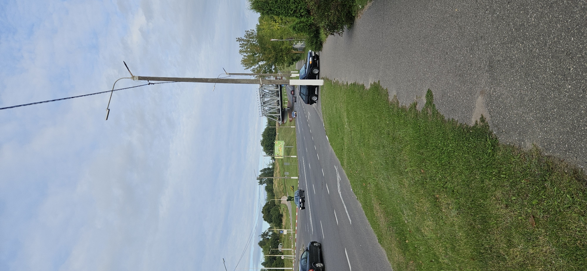 The image shows a street scene in Vitsebsk, Belarus. A paved sidewalk runs along the right side of the frame, bordered by a strip of green grass. Across the street, there are multiple lanes of asphalt road with white lane markings. A black sedan is parked on the side of the road next to the sidewalk. In the background, a large metal railway bridge spans across the road. Several cars are visible on the road, moving in various directions. Trees and green grass cover the landscape beyond the road, with a slightly overcast sky above. The overall scene depicts a typical urban environment with infrastructure and natural elements.