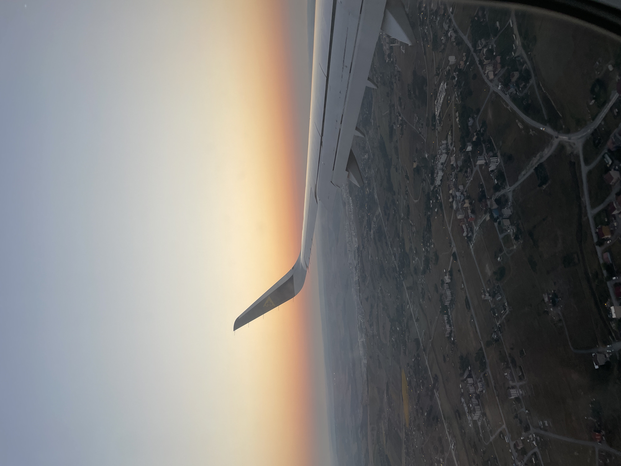 The image is a view from an airplane window, showing a wing and a landscape below at dusk. The sky exhibits a gradient of colors, transitioning from a pale blue at the top to soft orange and pink hues near the horizon, indicating a sunset. The airplane's wing, predominantly white with a dark grey logo on the wingtip, extends across the right and upper portions of the frame. Below the wing, a sprawling urban or semi-urban landscape is visible. The terrain appears to be hilly with scattered residential areas, featuring clusters of buildings, roads, and open fields. The lighting suggests it is either early morning or late evening, with the sun low on the horizon. The area appears to be a developed region, with visible infrastructure like roads connecting various settlements. The image does not contain any discernible people or specific landmarks that would pinpoint it to Beykoz, Turkey.