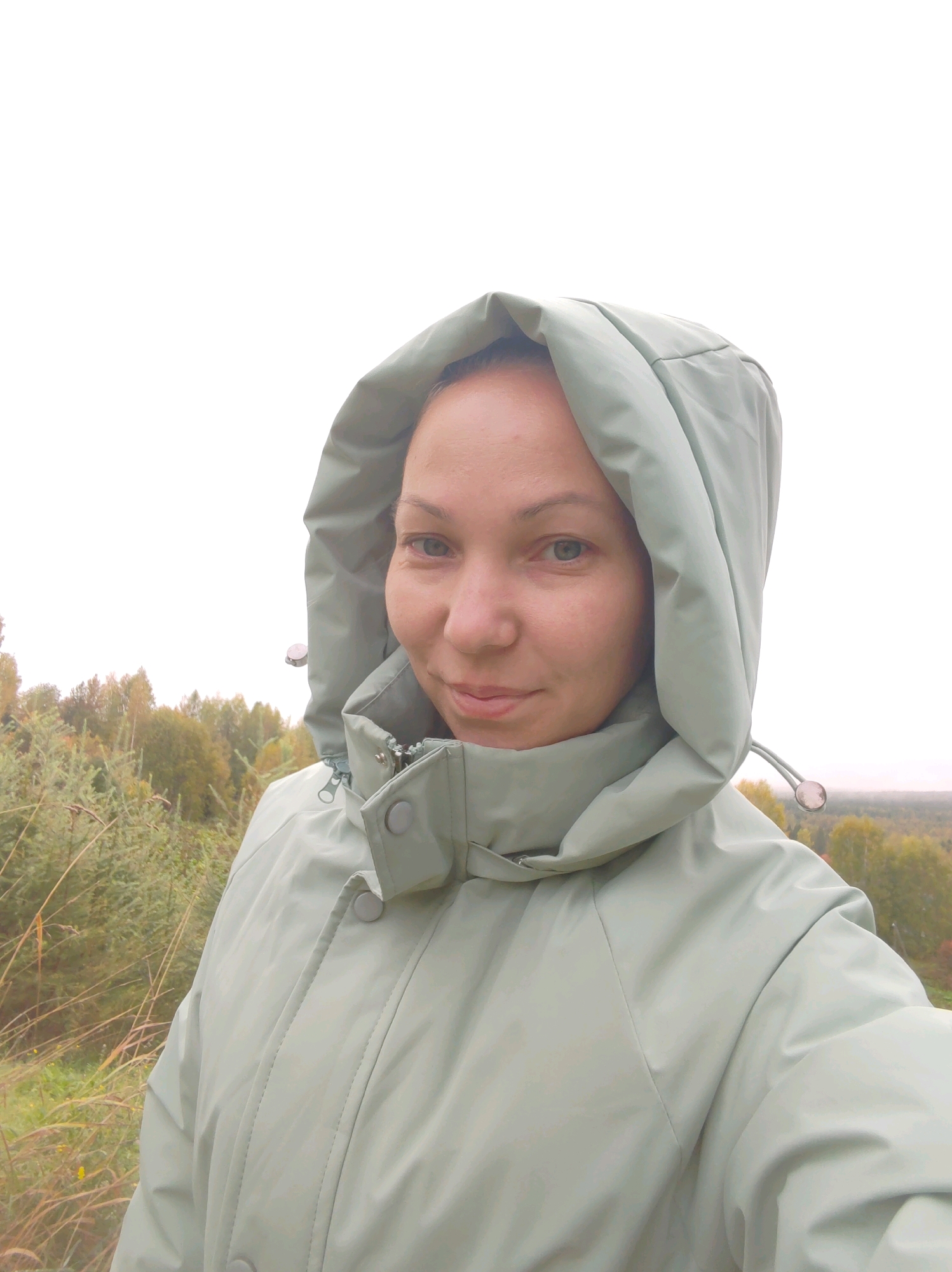 A medium-shot, selfie-style portrait captures a woman from the chest up. She is wearing a light green hooded jacket, with the hood up, framing her face. The woman has fair skin, light blue eyes, and is looking directly at the camera with a slight smile. The background shows a landscape of autumn trees and vegetation under an overcast sky. The exact location within the city of Чухлэм, Russia, is not discernible from the visual information provided in the image.