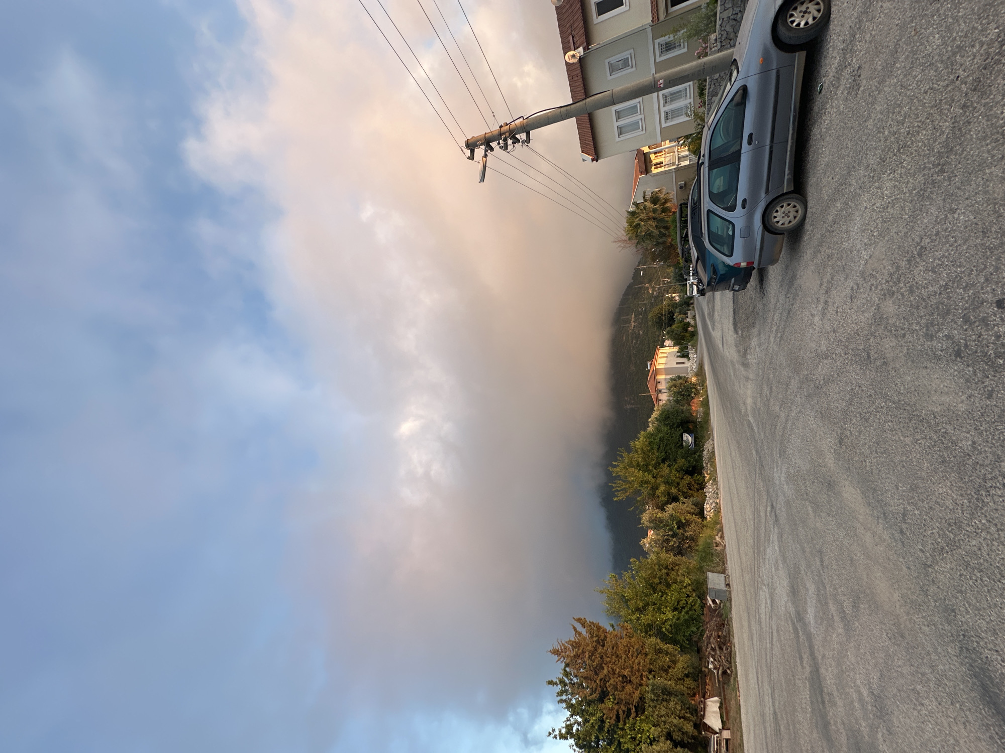 This image, captured in Fethiye, Turkey, depicts a residential street scene at either dawn or dusk, characterized by soft, warm light illuminating low-hanging cloud formations. The sky transitions from clear blue in the upper left to lighter shades and a dominant mass of grey, white, and peachy-pink clouds in the center and right, obscuring the upper slopes of a distant, tree-covered mountain.
The foreground features a wide, unpaved or poorly paved grey road, curving slightly towards the left. On the left side of the road, a cluster of various trees and shrubs separates the thoroughfare from an area containing discarded wood, debris, and a small metal bin. On the right side of the road, two cars are parked: a light blue/grey station wagon (possibly a Fiat Palio Weekend) in the foreground, and a darker-colored SUV behind it.
Behind the parked cars and alongside the road, several two-story houses with light-colored walls and reddish-brown tiled roofs are visible, consistent with local architecture. A prominent grey concrete utility pole stands on the right, supporting multiple overhead power lines and a streetlamp fixture, partially obscuring the view of the sky and buildings. Various trees, including olive trees and a palm tree, are interspersed throughout the residential area. No people or animals are visible in the scene.
