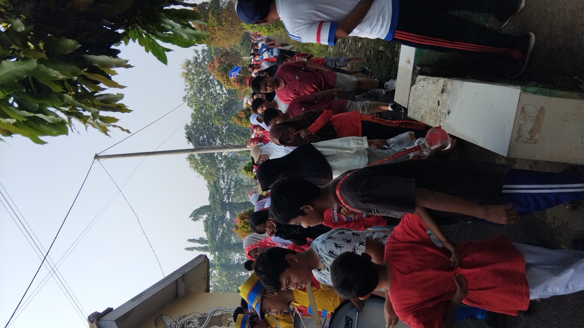 The image captures a street scene in Poso, Indonesia, likely during a procession or parade. A diverse group of people, primarily children and young adults, are walking in a line. Many of the children are dressed in casual clothing, while some are wearing uniforms or festive attire, suggesting a school or community event. A few individuals are holding musical instruments, specifically drums, indicating a marching band is present. The environment appears to be an outdoor urban setting with a road, trees, and buildings visible. The sky is clear and bright, suggesting a daytime event.