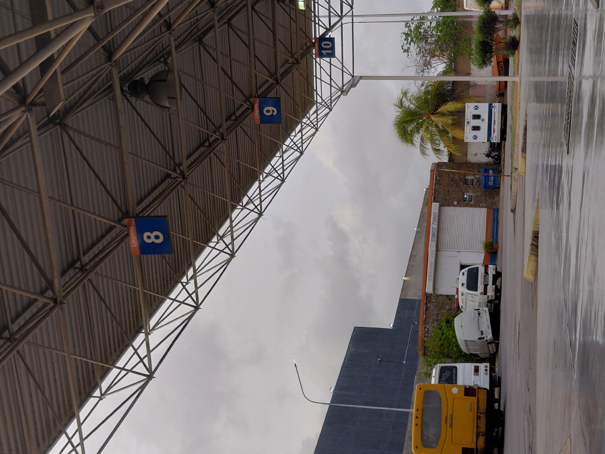 The image depicts an overcast day at a bus terminal in Juan Griego, Venezuela. A large, corrugated metal roof structure supported by a steel frame extends over a paved area. Red and blue signs with white numbers are visible hanging from the underside of the roof: "Llegada 8" (Arrival 8), "Llegada 9" (Arrival 9), and "10" (partially obscured). To the left, a yellow bus with its rear facing the camera is parked. Next to it are two white vans, one with its back doors open, and another white ambulance-like vehicle. Further to the right, another white vehicle, possibly a bus or van, is visible. A building with a brick facade and a white section with a garage-style door is present in the background. Trees, including a palm tree, and dense foliage are scattered throughout the scene. The paved ground appears wet, suggesting recent rain. Streetlights are mounted on poles. The overall atmosphere is utilitarian and industrial.