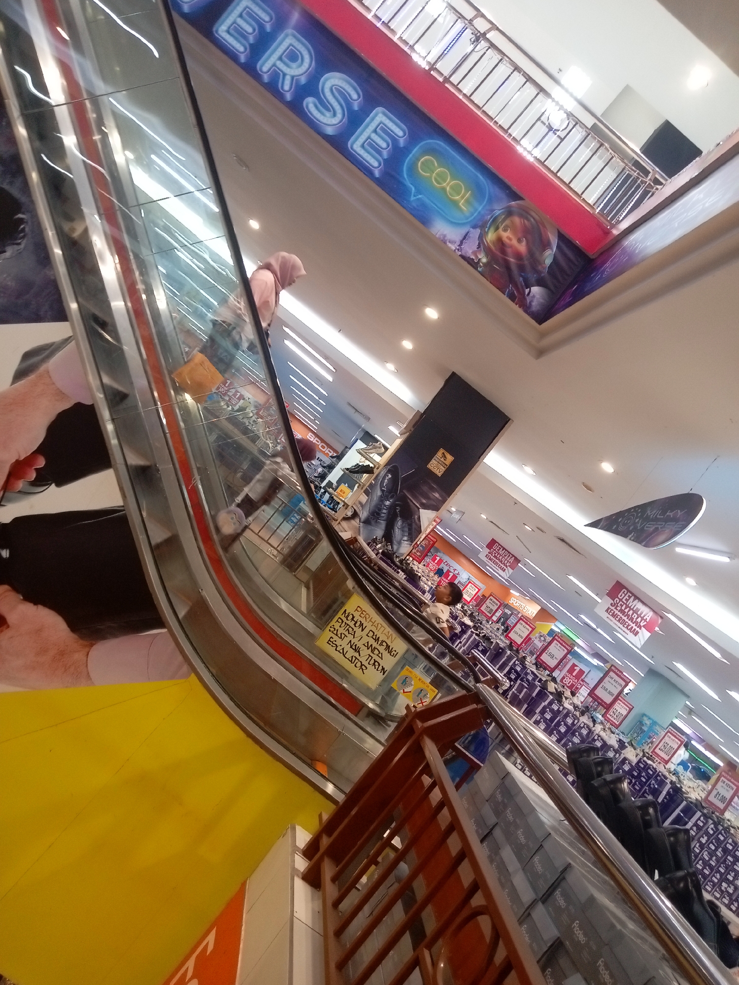 The image shows an interior scene of a shopping mall, likely in Batam, Indonesia, featuring an escalator. A person wearing a pink headscarf and a light-colored top is ascending the escalator. The escalator's metal railing and glass enclosure are visible, reflecting the surrounding environment. Above the escalator, a large banner with blue and red lettering is displayed, reading "UNIVERSE COOL" in stylized fonts. Another banner, featuring a cartoon character, is positioned below this. The mall's interior is illuminated by overhead lighting. Rows of merchandise, possibly shoes or clothing, are displayed on shelves in the background, with price tags visible. A CCTV sign is also present on a dark panel in the background. The overall environment appears to be a retail space within a commercial building.
