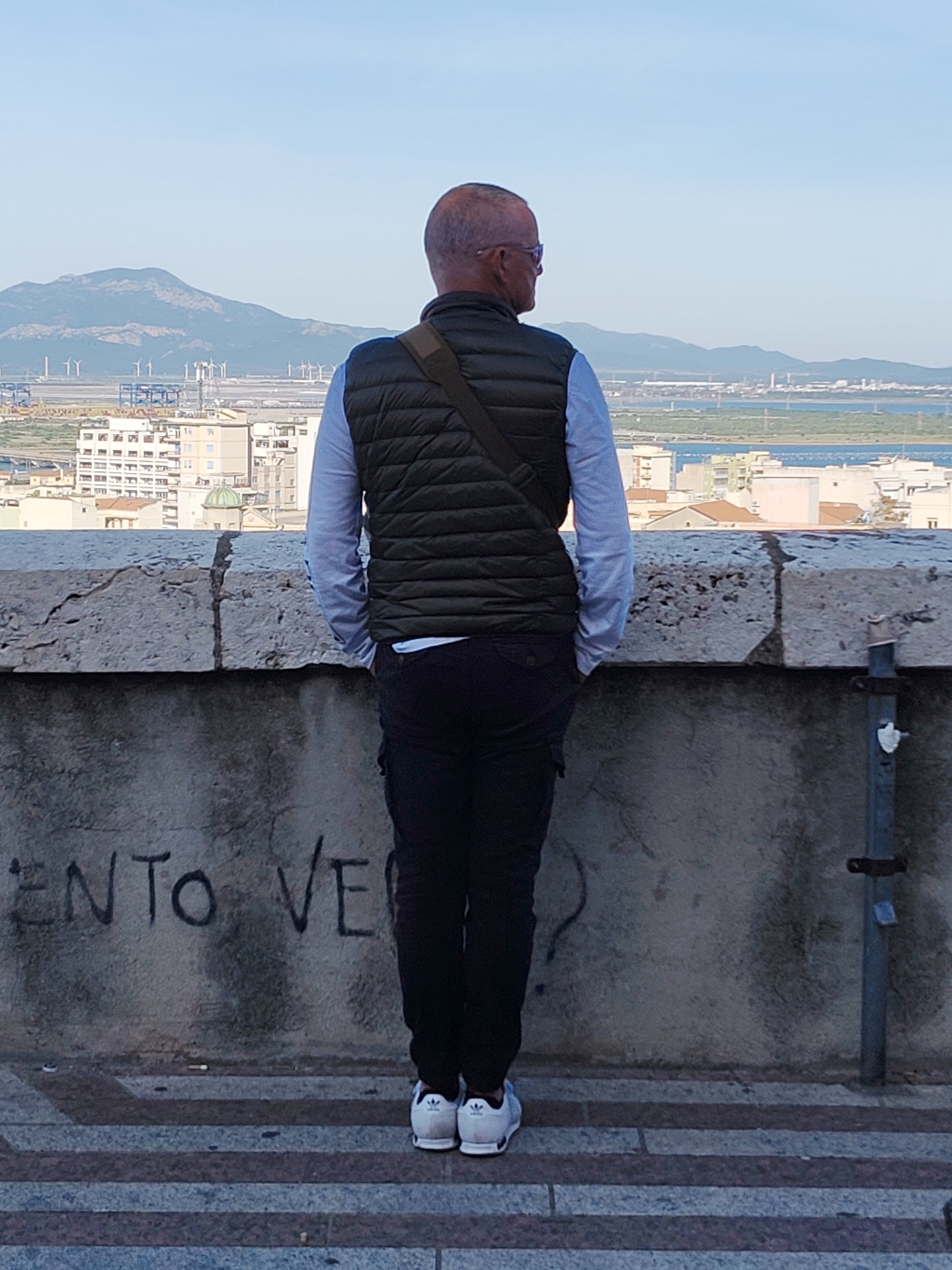 An outdoor scene in Cagliari, Italy, features a man standing with his back to the camera, gazing out over the city. He is positioned on a paved area next to a stone barrier or wall, behind which a cityscape is visible. In the background, there are mountains and possibly a bay area with what appear to be wind turbines or industrial installations on the horizon. The man is wearing a light-colored long-sleeved shirt, a dark olive-green vest, dark trousers, and white sneakers with dark accents. He also appears to be wearing glasses and has a bald head. The wall behind him has some graffiti written on it. The scene appears to be taken on a clear day with good visibility.