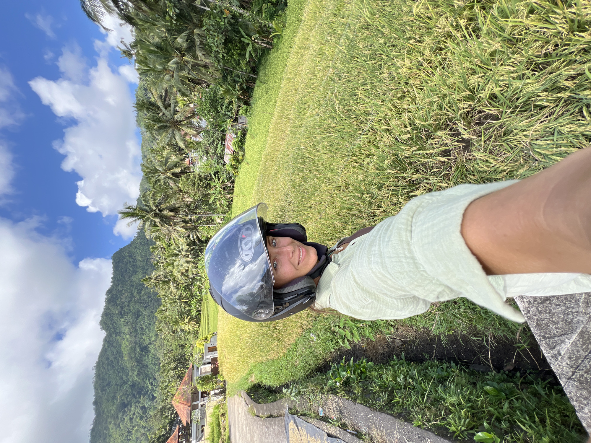 A selfie photograph shows a woman wearing a black KYT motorcycle helmet, her face turned toward the camera with a smile. She appears to be positioned near a rice field in the village of Sidemen, Indonesia. The photograph captures a partial view of a rice field, transitioning to lush green vegetation near a small cement ditch. In the background, a mountain covered in dense trees is visible under a blue sky with scattered clouds. Some houses with red tile roofs can be seen along the bottom of the mountain. The perspective indicates the image was taken from a lower angle, as if she is on a slightly elevated surface. A portion of the woman's light green shirt and arm are also visible in the frame.