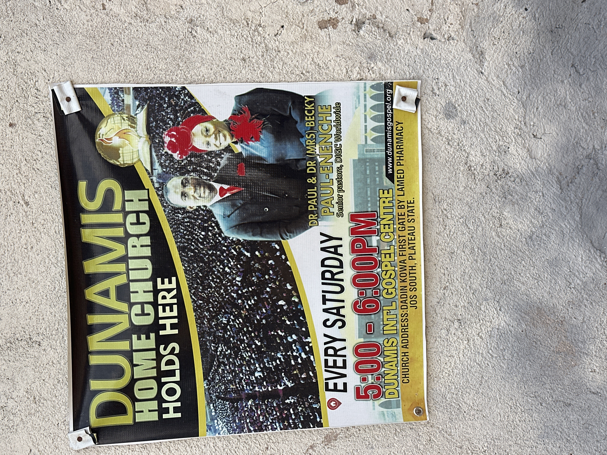 The image shows a poster advertising "DUNAMIS HOME CHURCH HOLDS HERE." The poster features images of Dr. Paul and Dr. (Mrs) Becky Paul-Enenche, identified as senior pastors of DIGCC Worldwide. Below their images, the poster states that services are held "EVERY SATURDAY" from "5:00 - 6:00PM" at "DUNAMIS INTL GOSPEL CENTRE." The church address is listed as "DADIN KOWA FIRST GATE BY LAMED PHARMACY, JOS SOUTH, PLATEAU STATE." A website address, "www.dunamisgospel.org," is also present. The poster is affixed to a rough, textured concrete wall with visible staples. The lighting suggests an outdoor setting with natural light. The overall scene depicts a public announcement in an urban or semi-urban environment.
