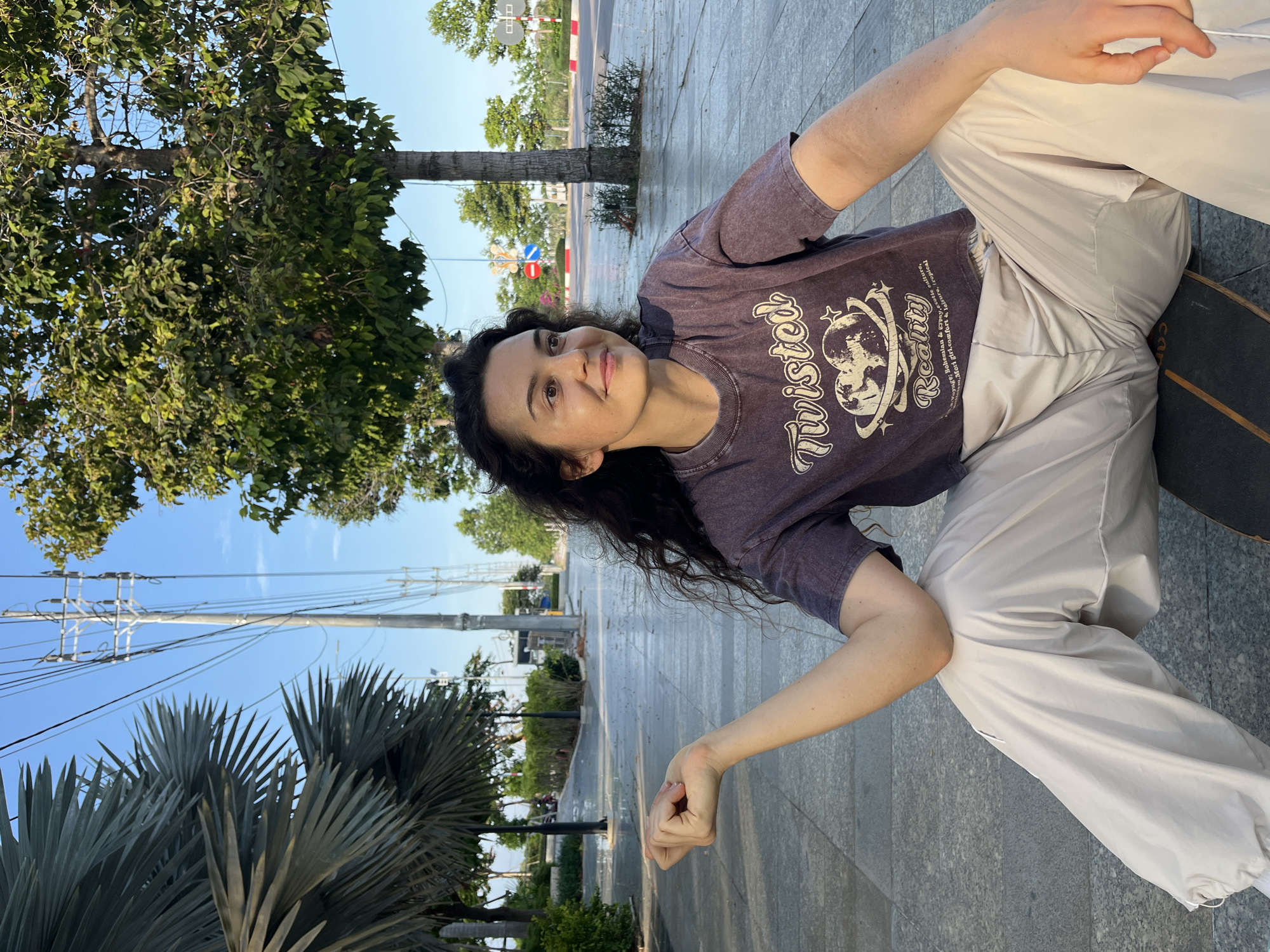 In this full-body, vertical shot, a woman with long, dark, wavy hair sits on a skateboard on a paved walkway. She is wearing a dark purple, slightly distressed t-shirt with a graphic print that includes the text "Twisted Reality" and imagery of planets and stars. Her pants are a light beige color with a loose fit. The woman is looking towards the camera with a slight smile.
The setting appears to be an outdoor urban environment. To the left of the woman are trees, including a large leafy tree and a palm tree with large fronds. Behind her, a road with traffic is visible, along with utility poles and wires stretching across the bright blue sky. The sun appears to be low in the sky, casting long shadows, suggesting it is either early morning or late afternoon. The overall mood is casual and relaxed, with the woman enjoying a moment outdoors. The text on her shirt, "Twisted Reality Voyage: Bohemian & Cyphy, exotic culture, Mori giri.comfort & leisure, tropical" is a notable detail, suggesting themes of travel, culture, and comfort.