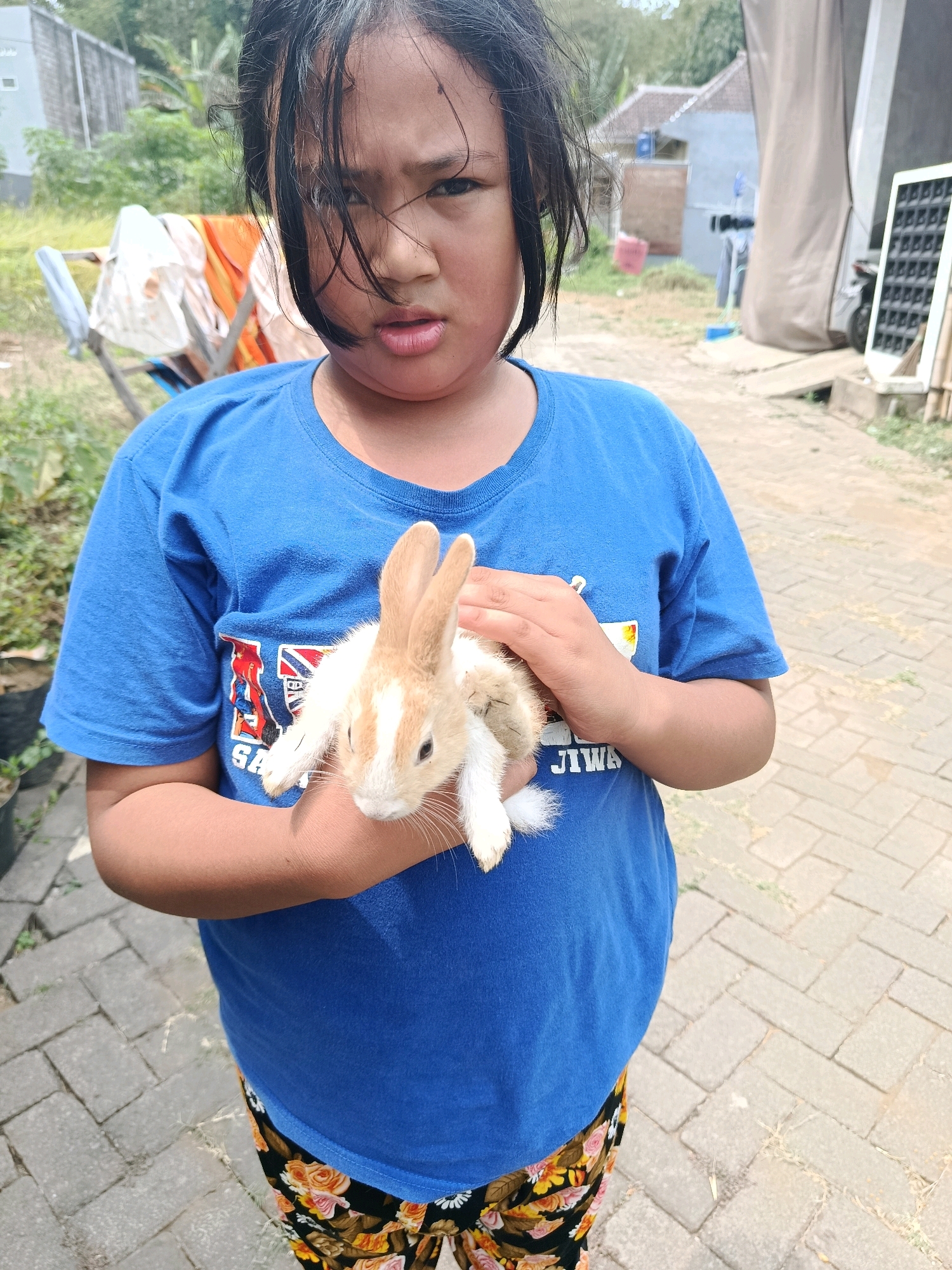 A young girl with dark hair stands outdoors in what appears to be a residential area in Bululawang, Indonesia. She is wearing a blue t-shirt with a graphic print and patterned leggings. The girl is holding a small, light-brown and white rabbit in her arms, cradling it against her chest. Her expression is neutral, with her eyes looking directly at the camera. In the background, there are houses, laundry drying on a rack, and some greenery. The ground is paved with light-colored bricks. The overall lighting suggests it is daytime.
