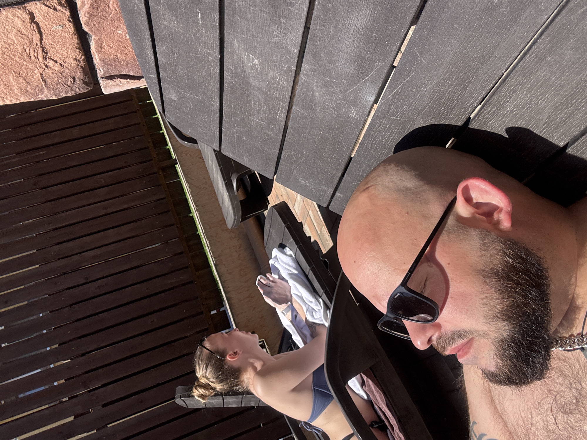 The image captures two individuals on an outdoor patio or deck constructed of dark wood. A woman, wearing a light blue bikini, reclines on a chair near a white towel. A man is situated closer to the camera. He has a shaved head and a full beard, and he is wearing sunglasses. He appears to be the subject of the selfie. The outdoor setting suggests a sunny day. In the background, there is a brick-like surface and some greenery. The image does not offer clear visual evidence to confirm the location in Krasnaya Polyana, Russia.