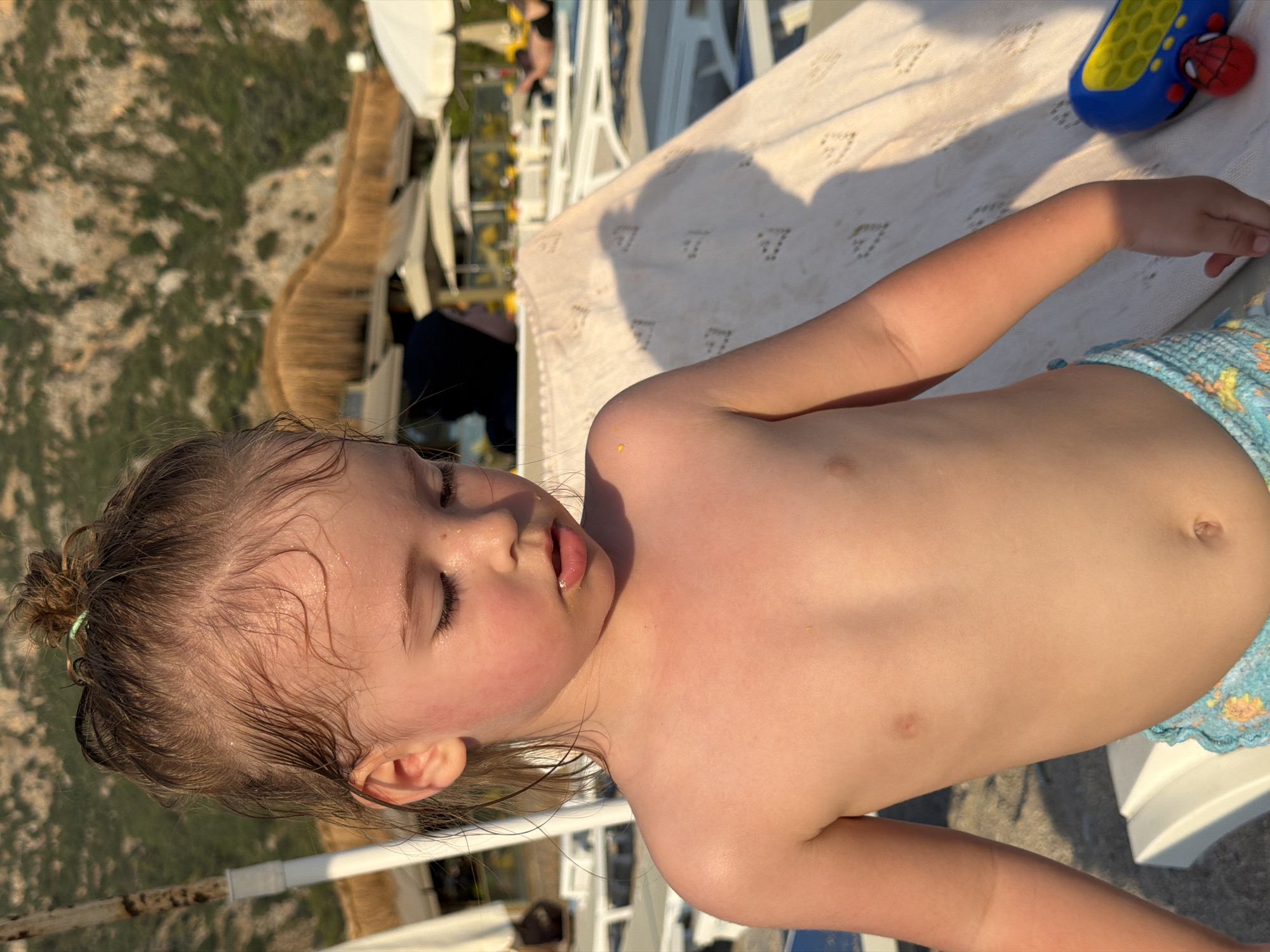 The image shows a young child lying down, possibly asleep, on a beach towel. The child is fair-skinned with wet, light brown hair pulled into a small bun. Their eyes are closed, and their mouth is slightly open. The child is wearing blue and yellow patterned swim trunks.
The scene appears to be outdoors, likely at a beach or poolside, given the presence of lounge chairs and a sandy or paved surface. In the background, there's a glimpse of a rocky hillside with sparse greenery, and what looks like a thatched umbrella structure. The lighting suggests it's either early morning or late afternoon, with warm sunlight casting shadows.
A blue and yellow toy with red accents, resembling a children's phone or electronic device with a Spiderman character, is visible in the upper right corner of the image. The overall mood is peaceful and relaxed, captured in a moment of quiet repose. The location is indicated as Fethiye, Turkey. There is no visible text in the image.