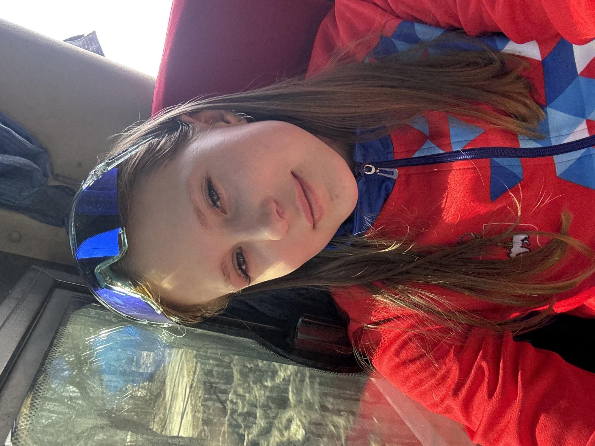 A young woman is sitting inside a vehicle, likely a car, and is the primary focus of the image. She is wearing ski goggles on her head and a red jacket with blue and white geometric patterns around the collar. She has long, brown hair. The vehicle's window is to her left. The seat is red, while other objects and shadows are visible in the periphery. The location in the city of Usolye, Russia is not discernible from the image.