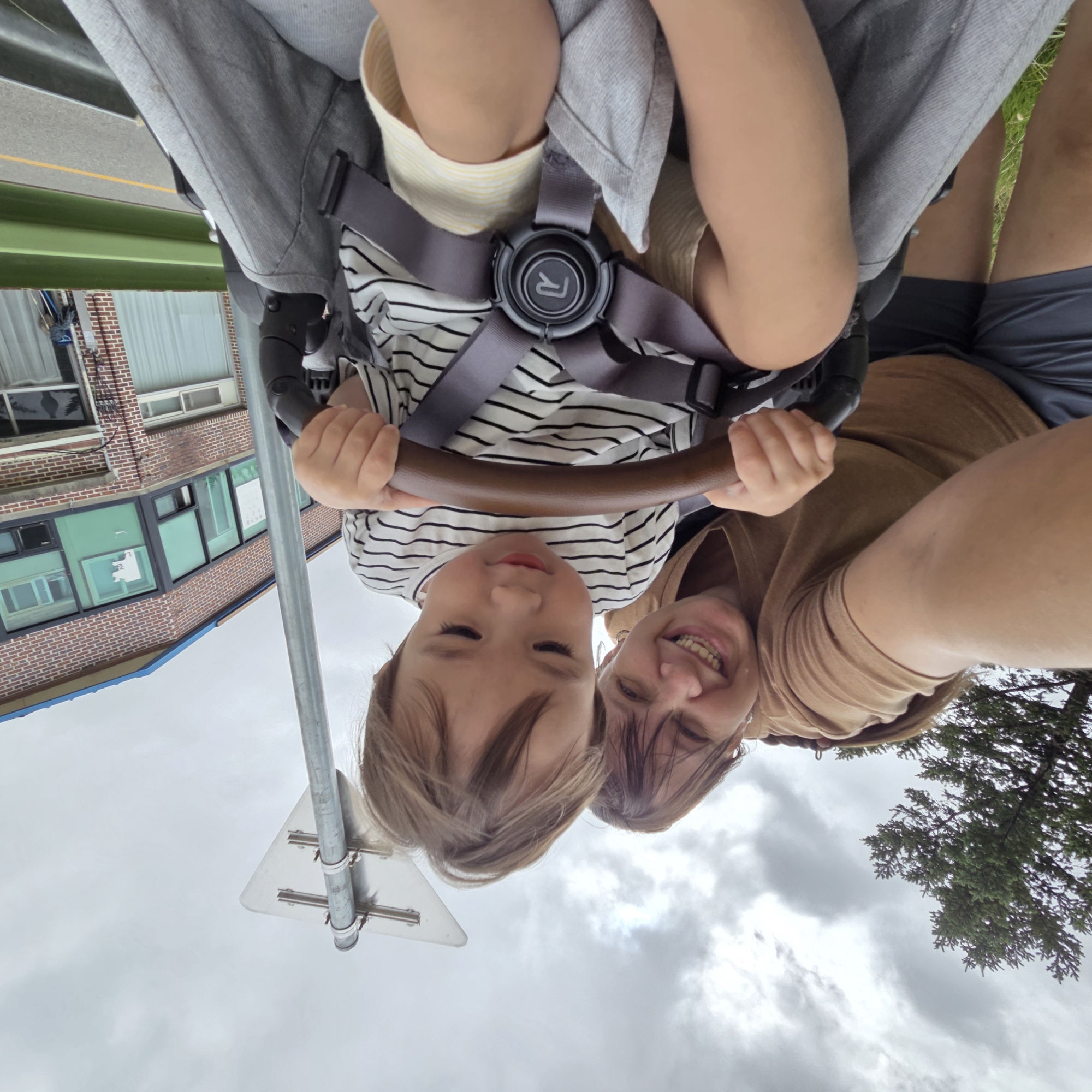 A woman and a young child are in a stroller, taken from a low angle. The woman, wearing a tan t-shirt, is on the left, smiling and holding the camera. The child, in a black and white striped shirt, is in the stroller, looking down at the stroller's handle. The background shows a building with brick walls and green-tinted windows, a traffic sign, a tree, and a cloudy sky. The location appears to be Dunnae-myeon, South Korea, based on the architectural style and presence of Korean text on a sign in the building's window. The scene suggests a casual outing, possibly a walk in a park or a visit to a local area.