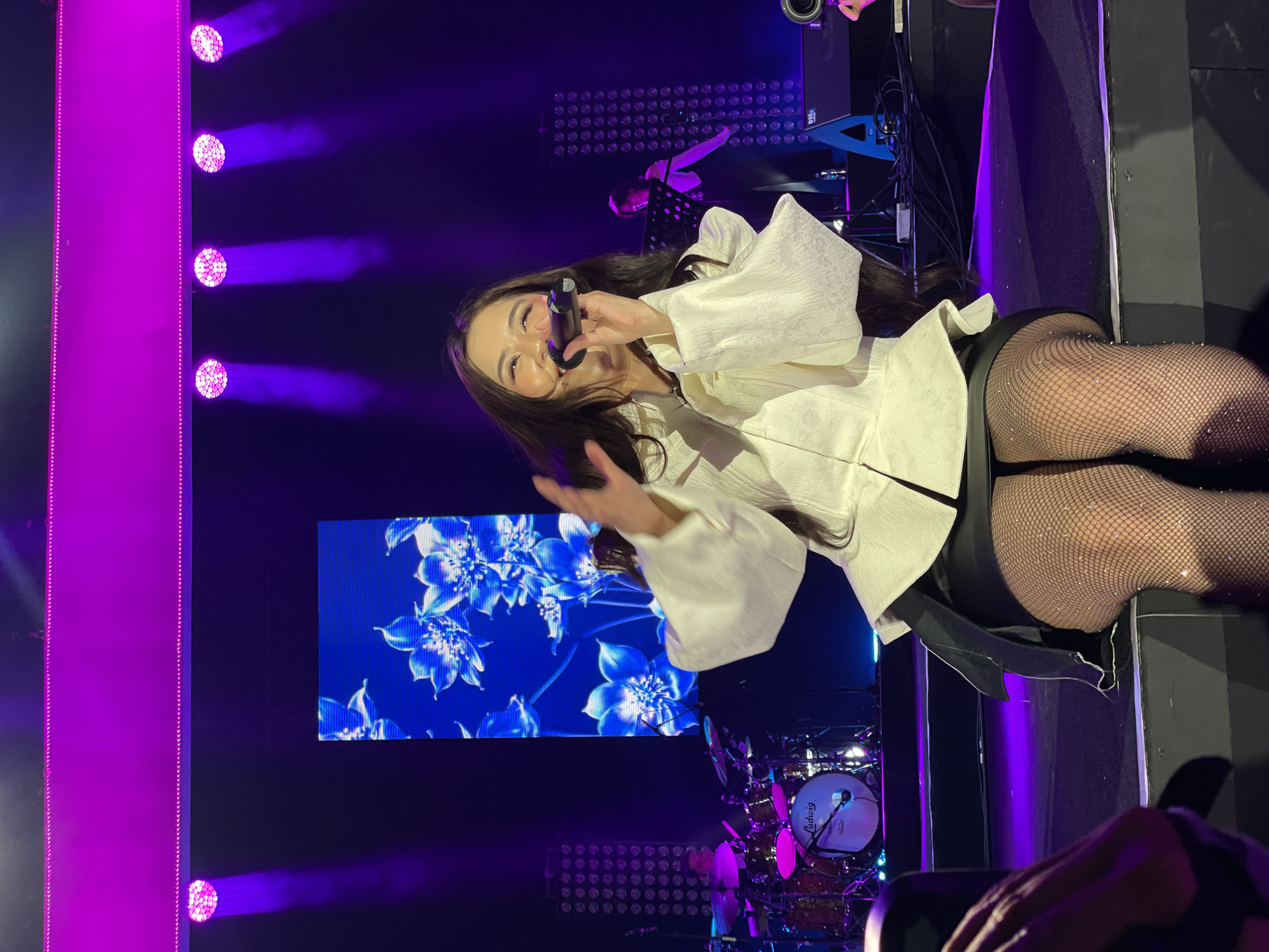 The image shows a female performer on a stage, likely during a concert or performance. She is holding a microphone and singing. She is wearing a white, long-sleeved shirt with wide sleeves, a black bottom, and fishnet stockings. Behind her, there is a large screen displaying blue and white flower graphics. Stage lighting creates a purple hue in the background. A drum set is visible at the bottom. Other people are present in the scene, including a musician and at least one person in the foreground holding a camera. The location is Salembaran, Indonesia.
