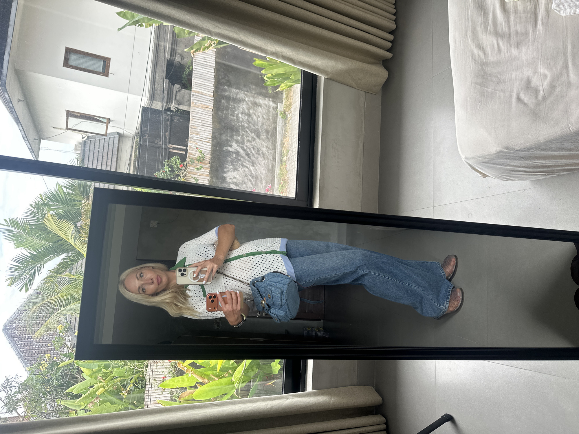 A blonde woman stands in front of a full-length mirror, taking a mirror selfie. She is wearing a white crochet top with green trim, blue denim jeans, and brown woven sandals. She is holding two phones in her right hand and a light blue quilted backpack hangs from her left shoulder. The mirror reflects a room with a bed covered in a light beige duvet and a window with beige curtains. Outside the window, palm trees and a building are visible, suggesting a tropical location. The scene appears to be in a private residence in North Kuta, Indonesia.
