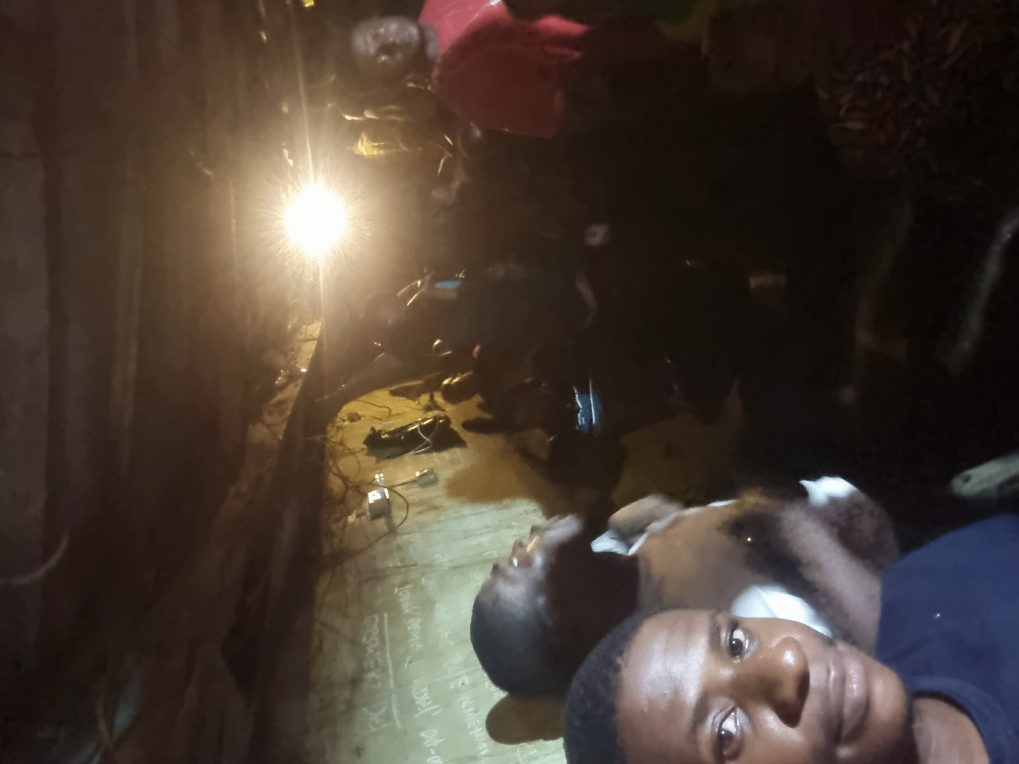 A dimly lit, indoor scene in Onitsha, Nigeria. A group of people are present, one individual is taking a selfie with a dark blue shirt, there is also an animal that is brown and white. A light source shines brightly in the background, illuminating a wall covered with chalk writings. Electrical wiring and plugs are visible along the wall.