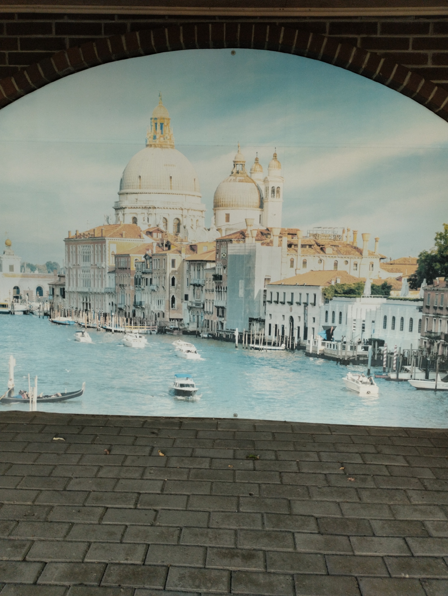 The image depicts a mural of Venice, Italy, displayed behind a brick archway and in front of a brick pathway. The mural shows a scenic view of the Grand Canal with several boats, including a gondola and motorboats, navigating the water. In the background, there are numerous historic buildings with distinct architectural styles, including domes and ornate facades, typical of Venetian architecture. The sky above is clear with some clouds, and the water of the canal is a vibrant blue. The brick archway framing the mural is constructed of dark red bricks. The foreground shows a pathway made of dark gray rectangular bricks laid in a staggered pattern. There are a few scattered leaves on the brick pathway. The image is a photograph taken from a slightly elevated perspective, looking down at the pathway and up at the mural. The mural is not located in Kostanay, Kazakhstan.