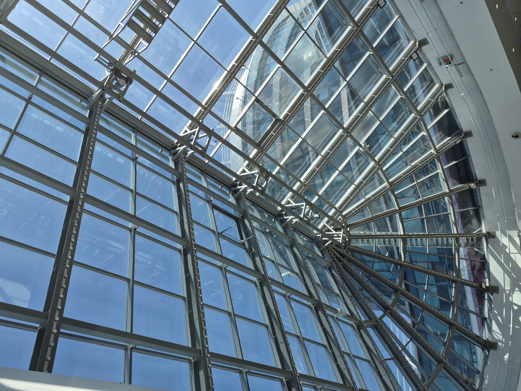 The image presents an upward view of an interior architectural structure, likely a glass ceiling or atrium, potentially located in Tobolsk, Russia. The structure consists of a grid of metal beams that support glass panels, allowing natural light to enter. The angle of the view gives the impression of looking up through a complex geometrical pattern. Through the glass panels, the sky is visible, as well as reflections of other buildings, implying an urban setting. The metal beams are constructed from a network of supporting steel, and the structure appears modern and well-maintained. At the far right, the edge of an interior wall suggests that the atrium is part of a larger building.