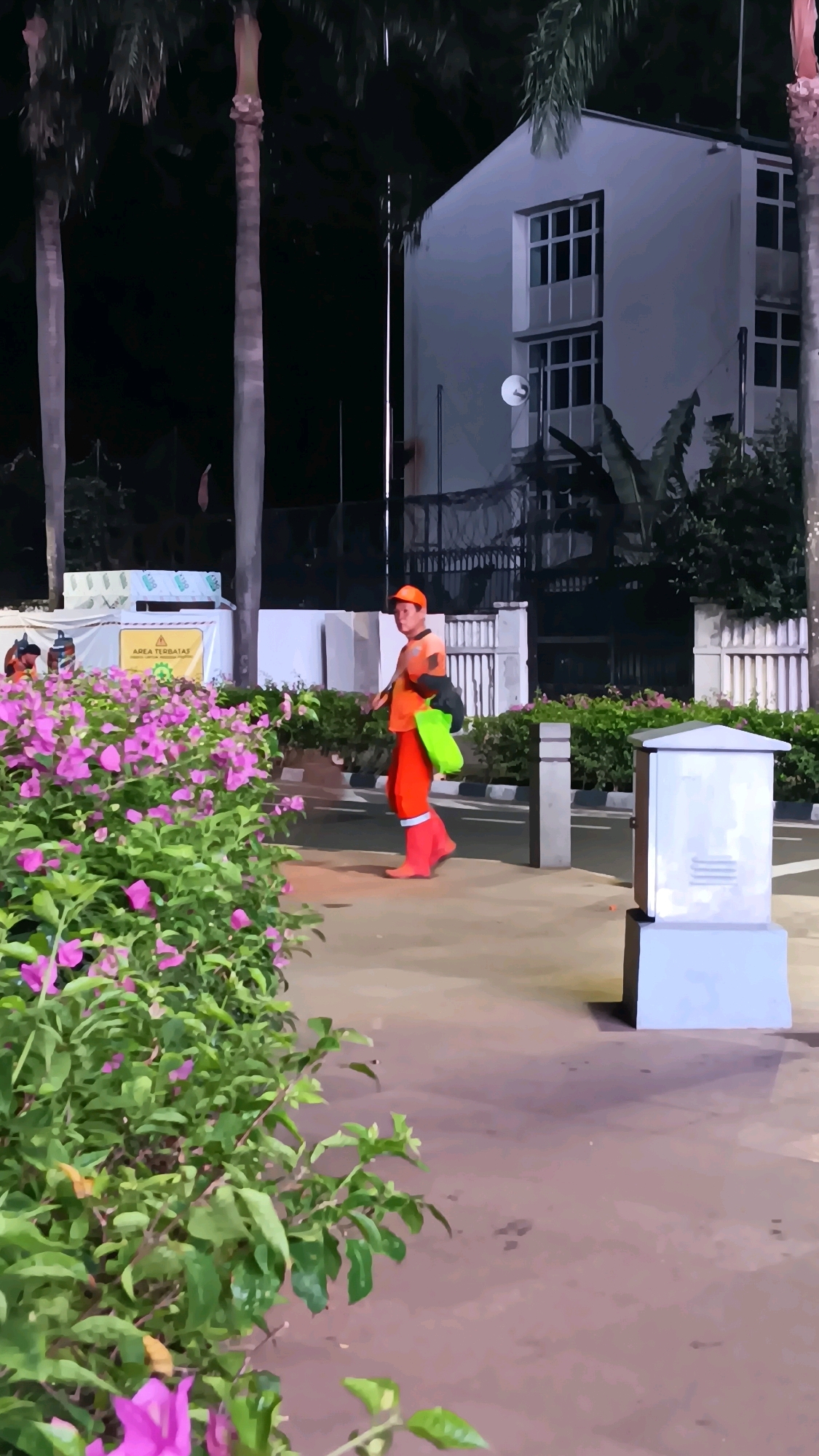 A lone sanitation worker, dressed in bright orange attire and matching rubber boots, walks across a paved area at night. He carries a green bag in his left hand and what appears to be a black garbage bag slung over his right shoulder. The worker is walking away from a white building with multiple windows, which is partially obscured by palm trees. In the foreground, a dense bush with vibrant pink flowers lines a pathway. The scene is illuminated by artificial lighting, creating shadows on the ground. A light gray electrical box is visible to the right of the worker. The location is Central Jakarta, Indonesia.