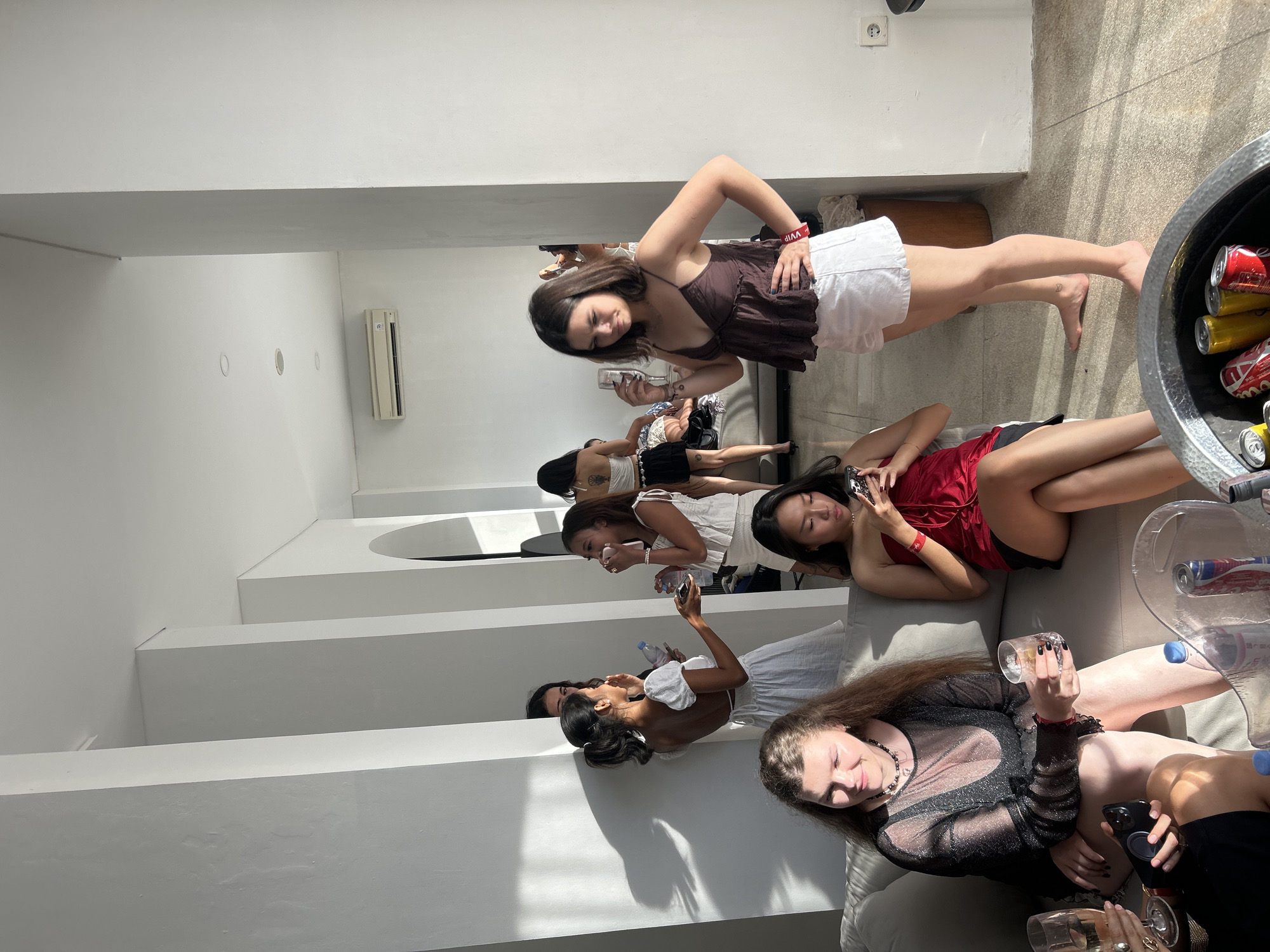 In a bright, modern interior space in North Kuta, Indonesia, a group of young women are gathered, appearing to be at a party or social event. The room features white walls, architectural columns, and a couch area. Sunlight streams in, creating shadows and illuminating the scene.
In the foreground, a woman with long, wavy brown hair is seated on a couch, wearing a sheer, sparkly black long-sleeved top and a black skirt. She holds a glass of amber liquid and a smartphone. Next to her, another woman in a vibrant red strapless top and black skirt is looking down at her phone. Partially visible to the left is a woman with blonde hair, wearing a black outfit, holding a champagne flute. A silver ice bucket with cans of drinks sits in the lower right corner.
In the mid-ground, standing and interacting, are several other women. One woman in a white dress with puffed sleeves is holding a bottle and speaking to another woman. Further back, a woman in a white top and skirt is holding a drink. Another woman in a white outfit is visible in the background.
In the right mid-ground, a young woman with short brown hair is standing, holding a glass and looking slightly away from the camera with a pensive expression. She wears a brown camisole and white shorts.
The overall atmosphere appears relaxed and social, with an emphasis on casual conversation and enjoyment of drinks.
