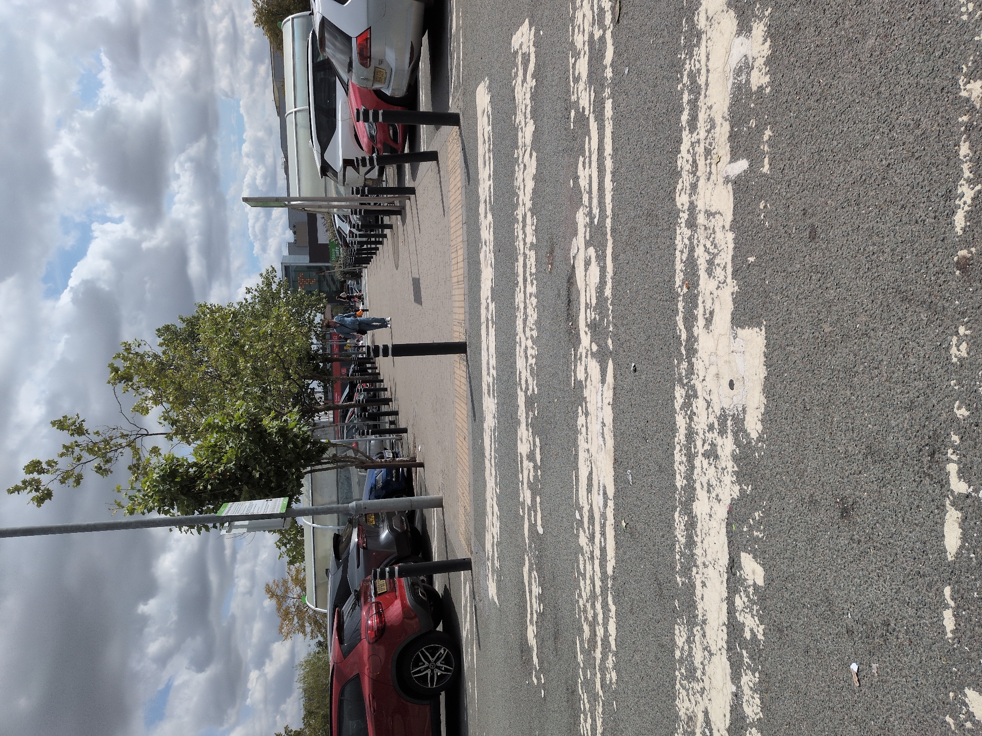 An exterior, eye-level shot captures a paved pedestrian crossing in a car park. The crossing's white stripes are faded and worn, indicating heavy use. Beyond the crossing, a paved walkway runs alongside parked cars, with bollards separating the walkway from the car park. A single person, dressed in a blue shirt and light-colored trousers, walks along the walkway, carrying a dark bag. In the background, trees with green foliage are visible, along with buildings and a partially obscured blue sign. The sky above is filled with a mix of white clouds and blue sky. The scene is likely in Telford, United Kingdom, given the specific context provided.